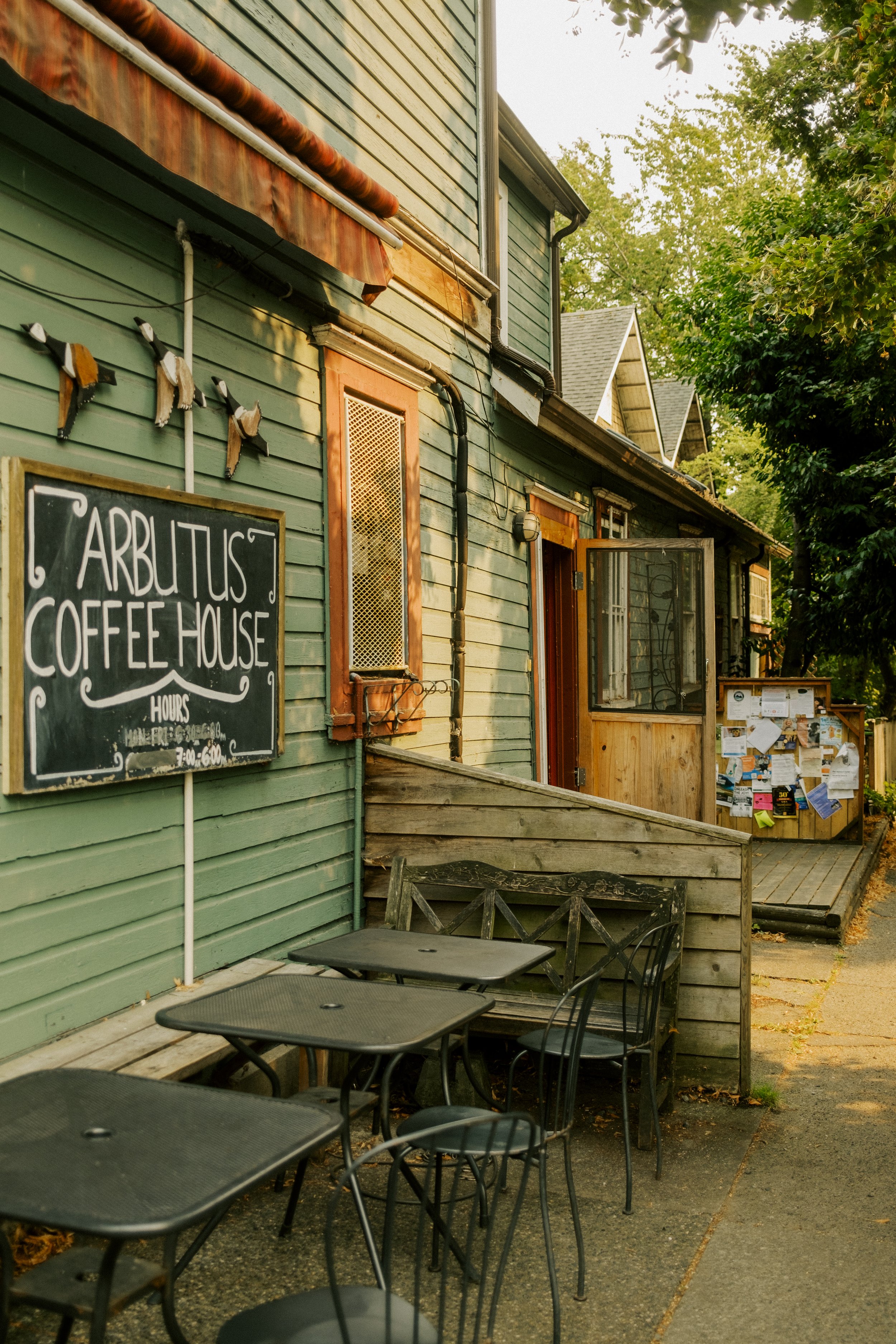 Exterior view of Arbutus Coffee House with outdoor seating, a chalkboard sign, and a bulletin board in a quaint, rustic building.