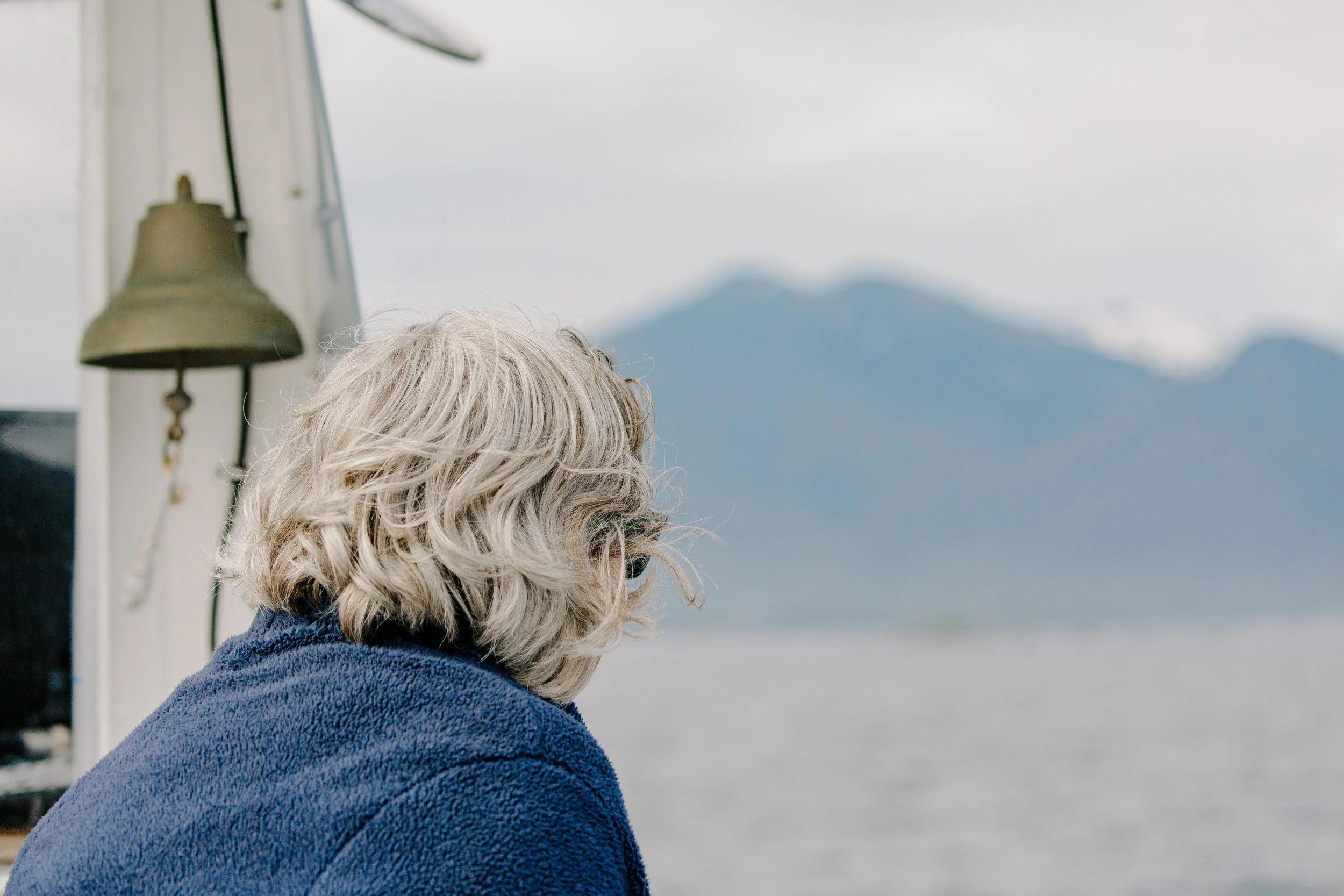 A person with gray hair wrapped in a blue towel, facing a body of water with mountains in the background.