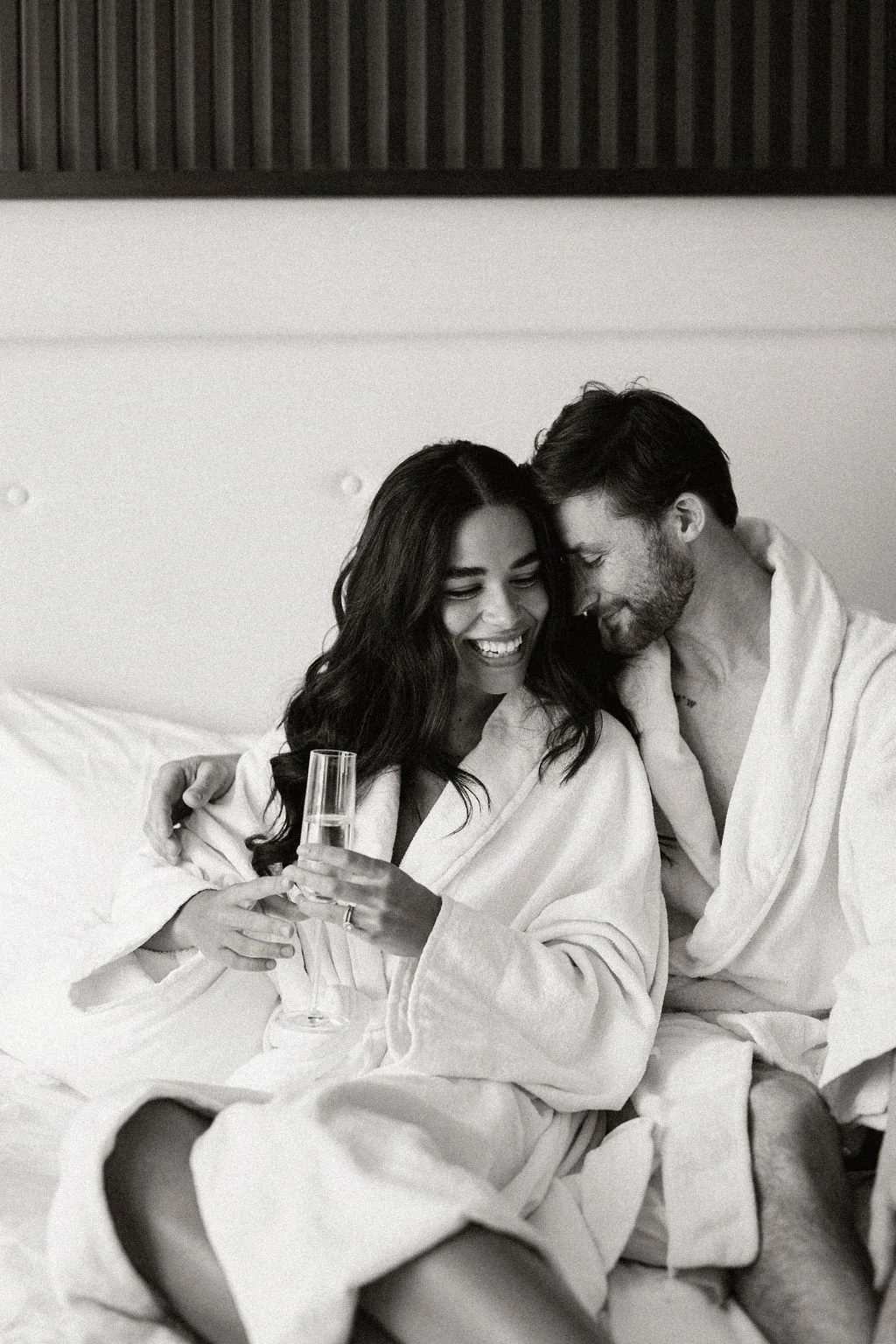 A happy couple in bathrobes sitting on a bed, with the woman holding a glass of champagne, sharing a joyful moment.