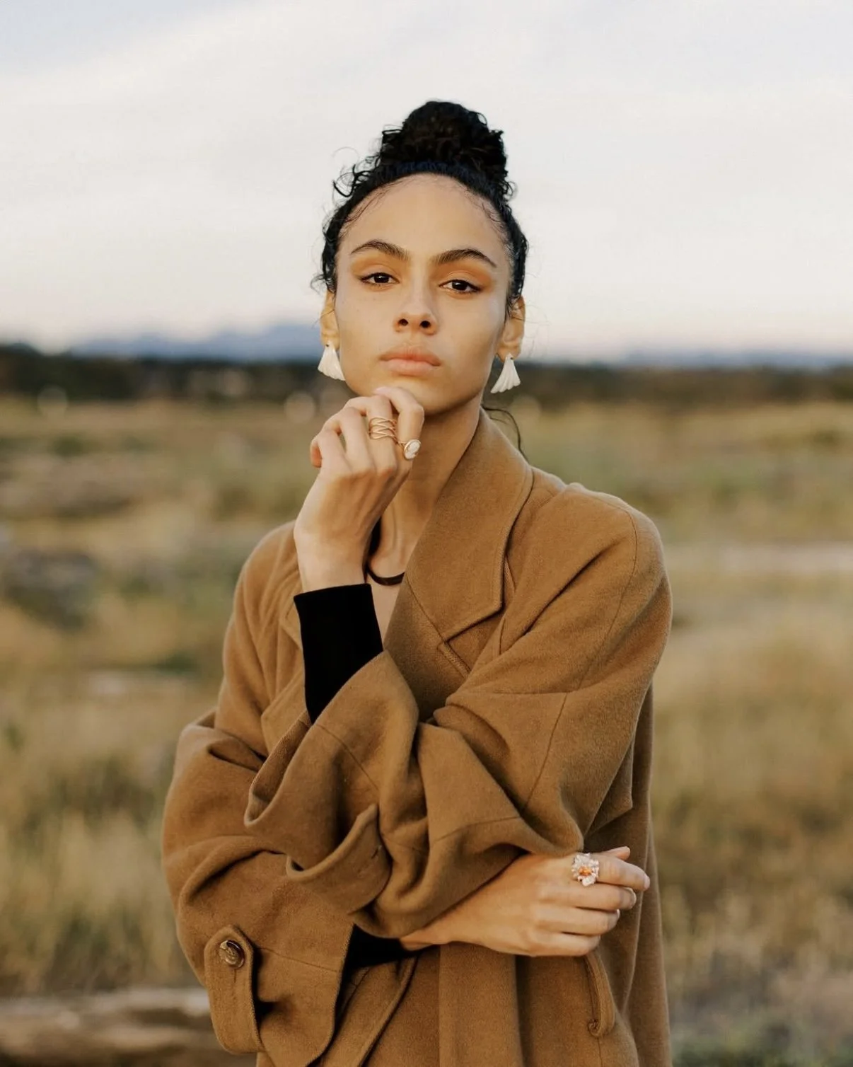 A young woman with dark hair tied up, wearing earrings, a brown coat, and rings, poses outdoors in a natural setting.