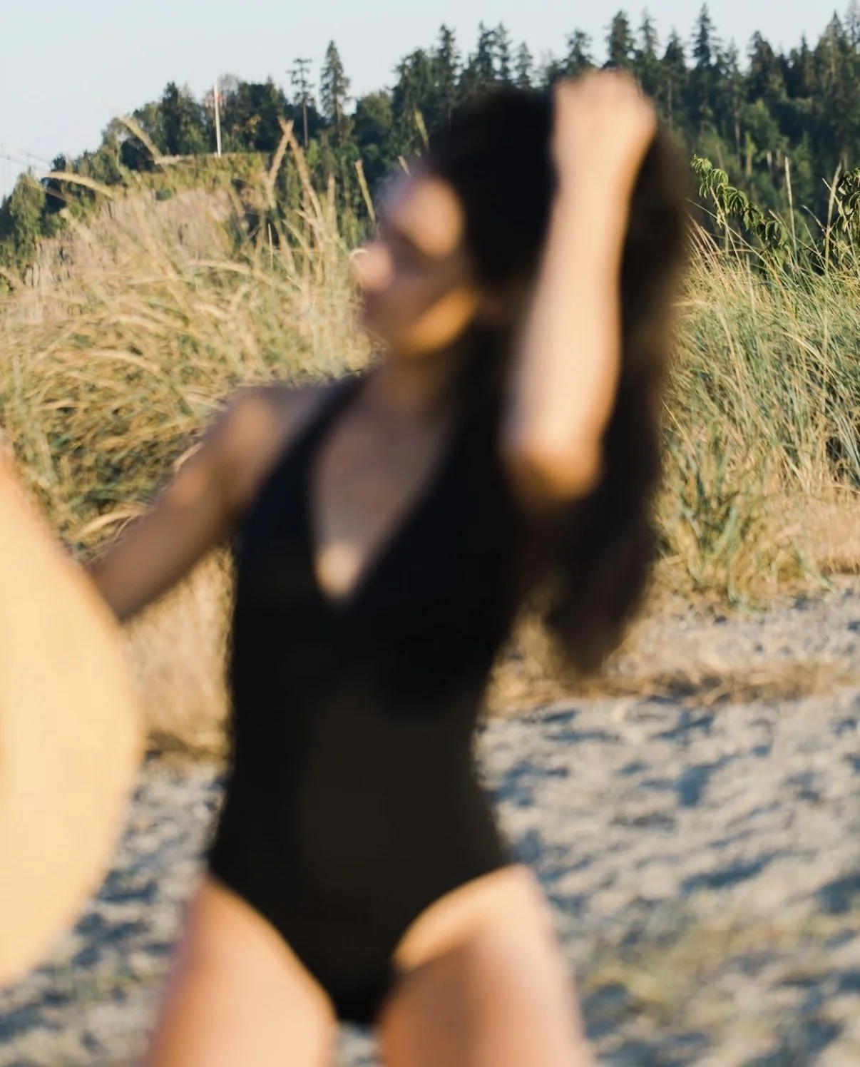 A woman in a black swimsuit taking a selfie on a beach with tall grass and trees in the background.
