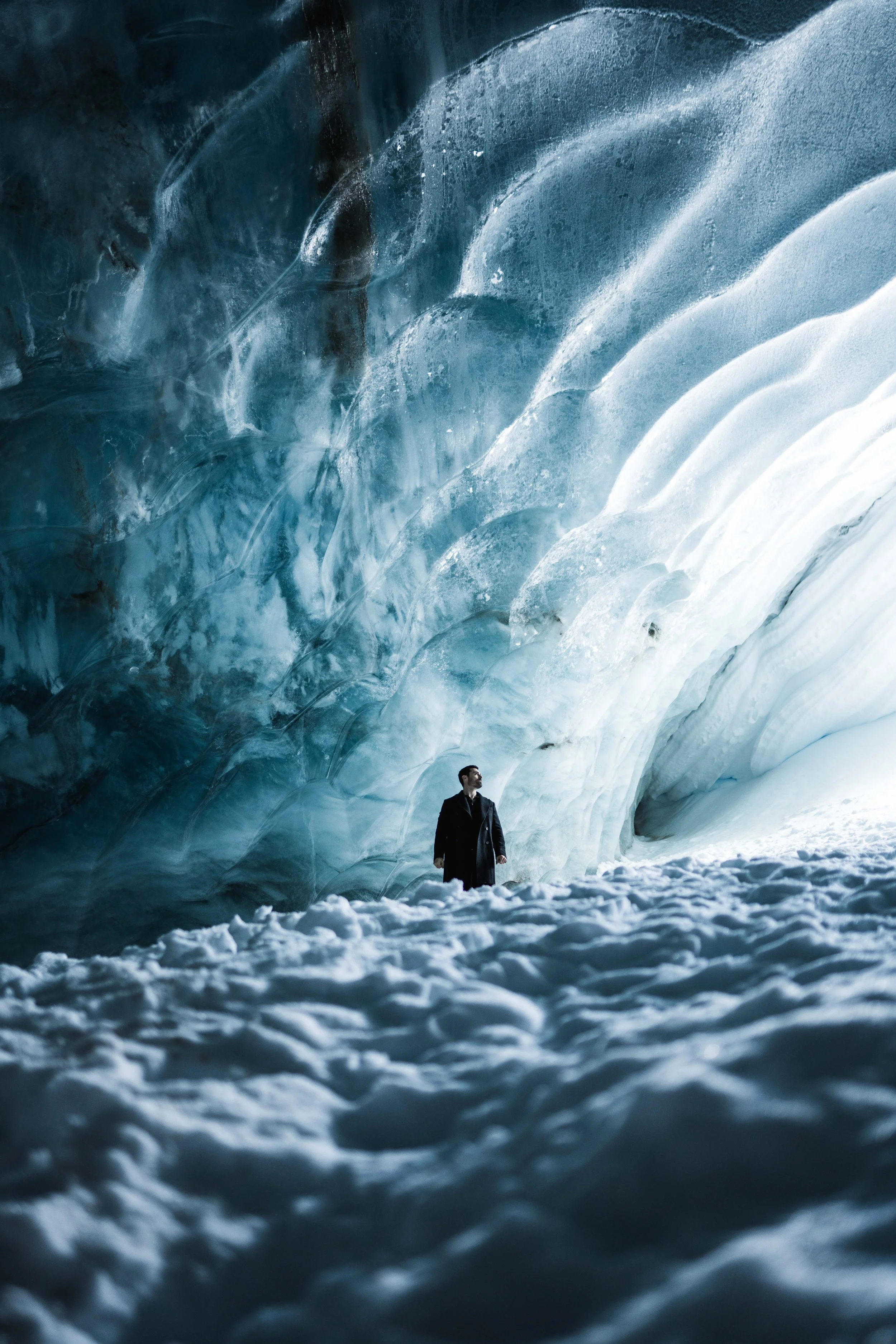 A man in a coat standing inside a large icy ice cave with blue and white ice formations.