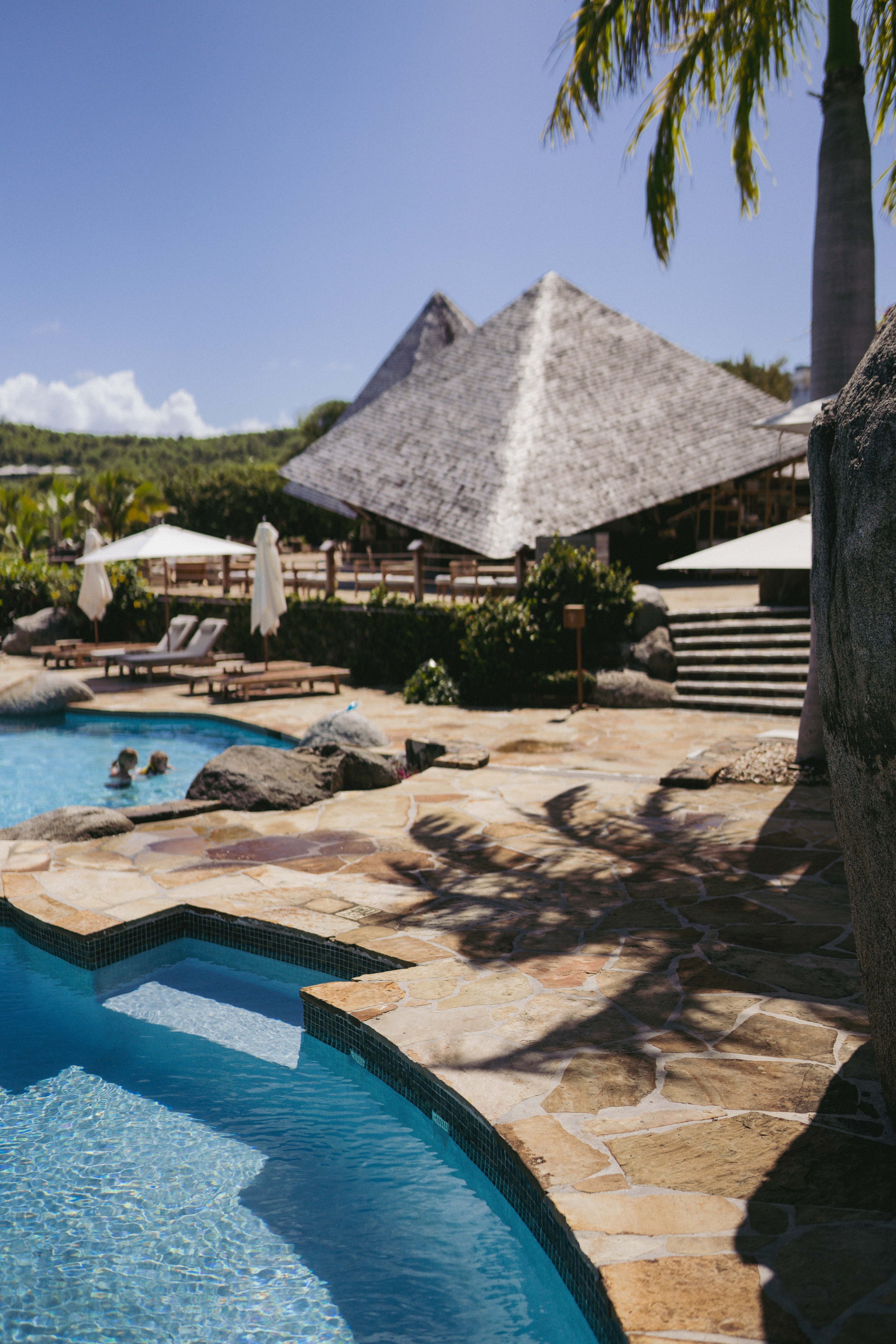 Resort pool area with lounge chairs, umbrellas, and a thatched-roof building under a clear blue sky, with palm trees and lush greenery in the background.