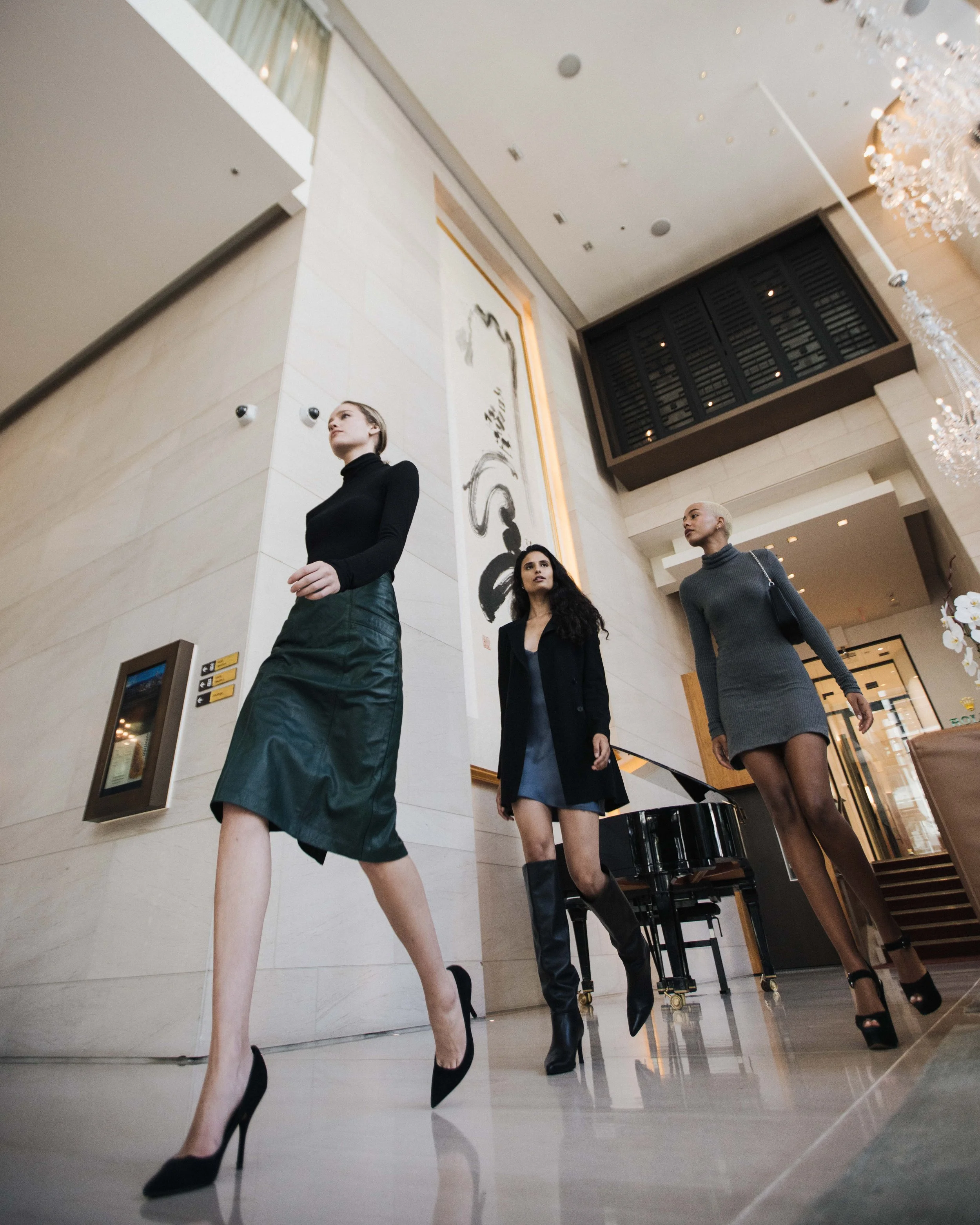 Three women walking through a hotel lobby, dressed in black and gray fashionable outfits, with a grand piano in the background.