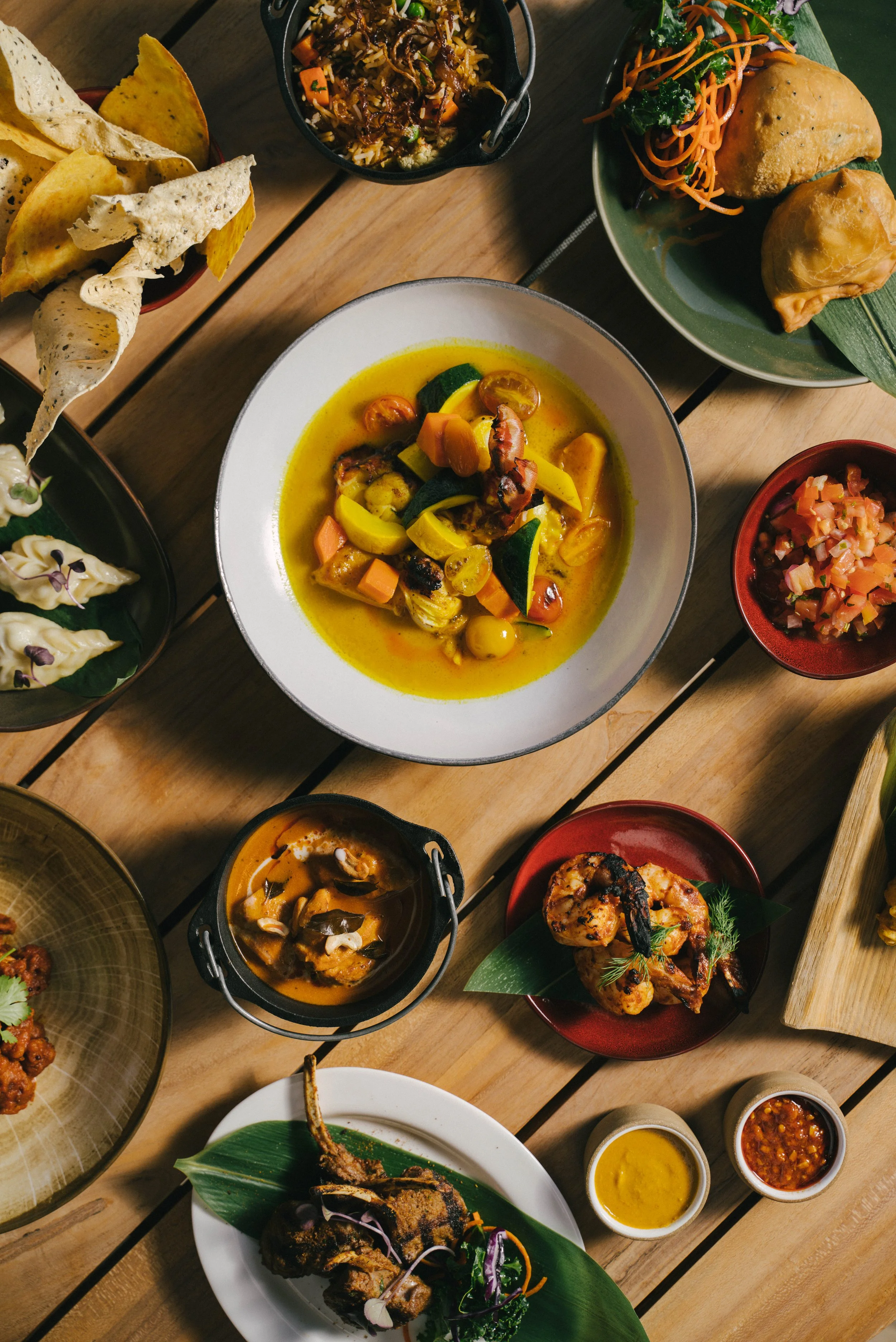 A variety of Indian dishes including curry, samosas, naan bread, and chutneys on a wooden table.