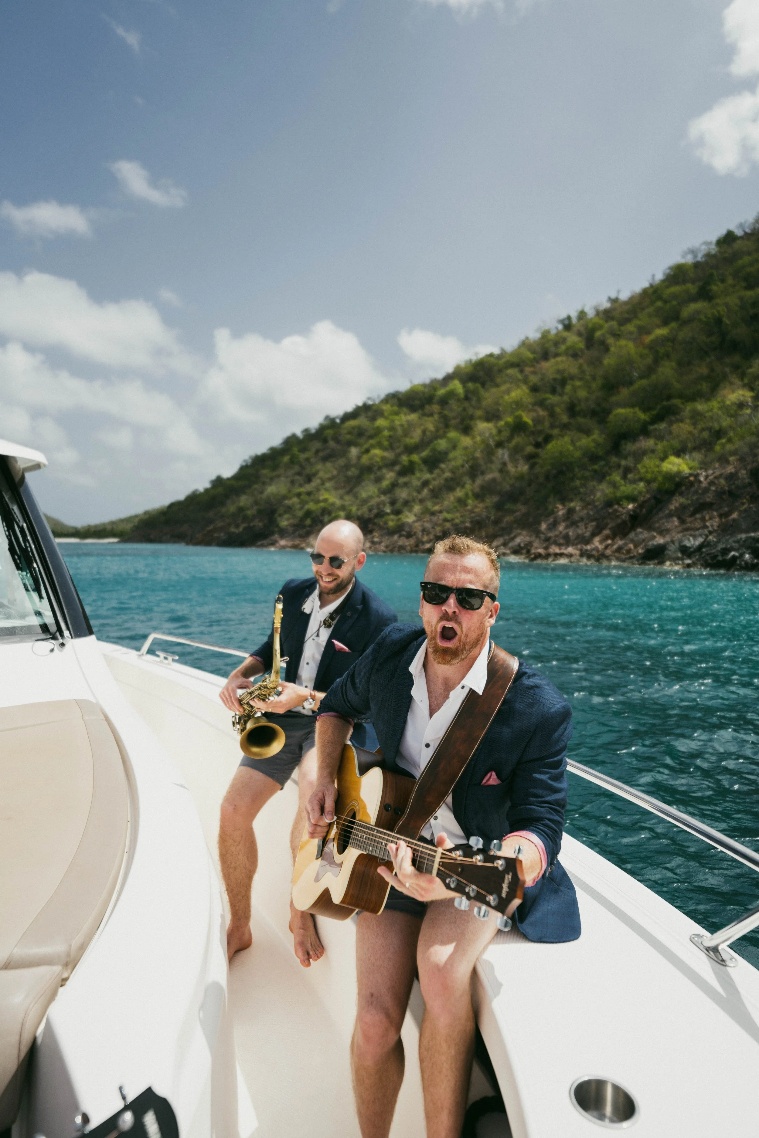 Two men in suits without pants playing musical instruments on a boat in the water, with green hills and blue sky in the background.