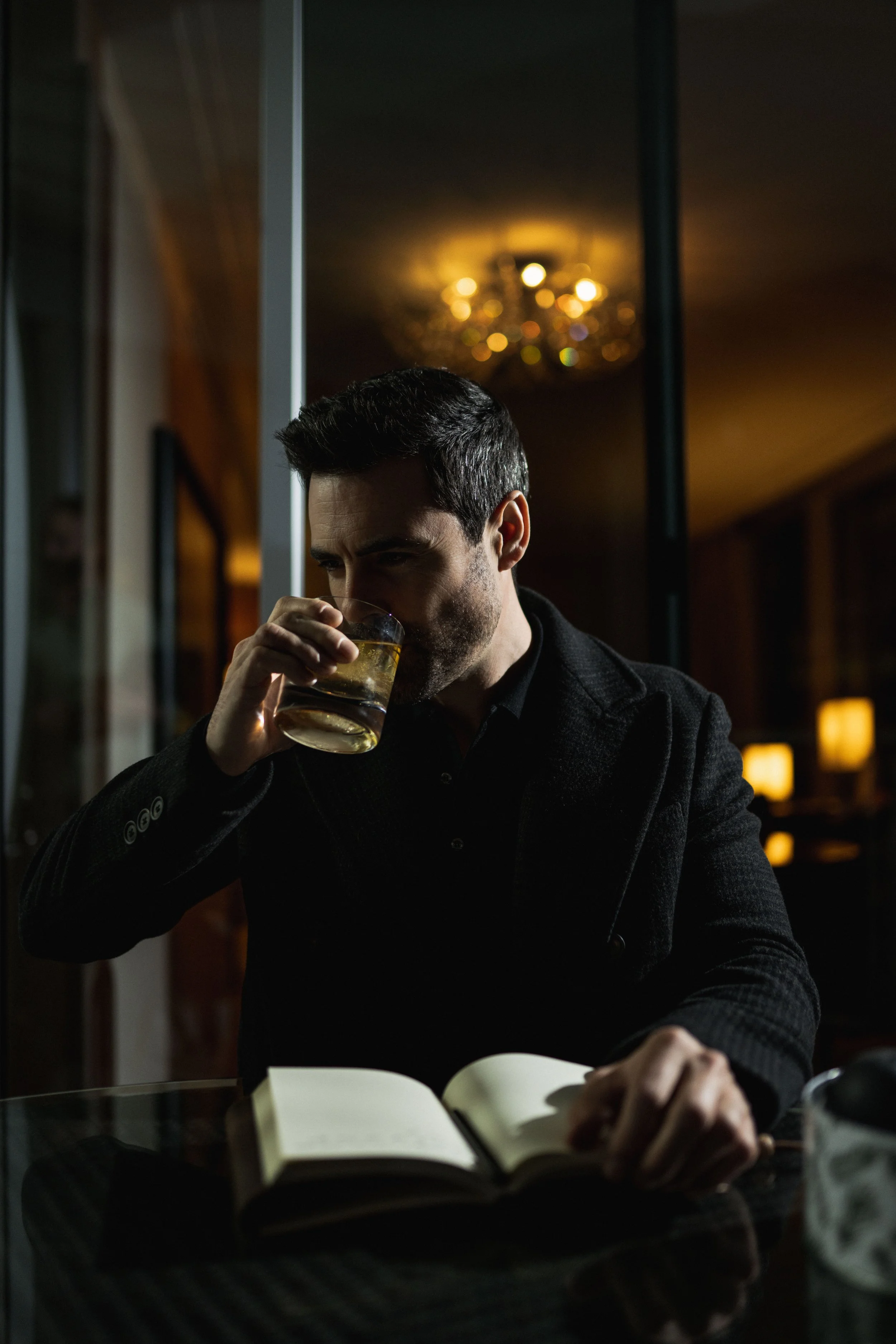 A man in a black blazer is sitting at a table, drinking a glass of whiskey, with an open book in front of him, in a dimly lit room with warm lighting.