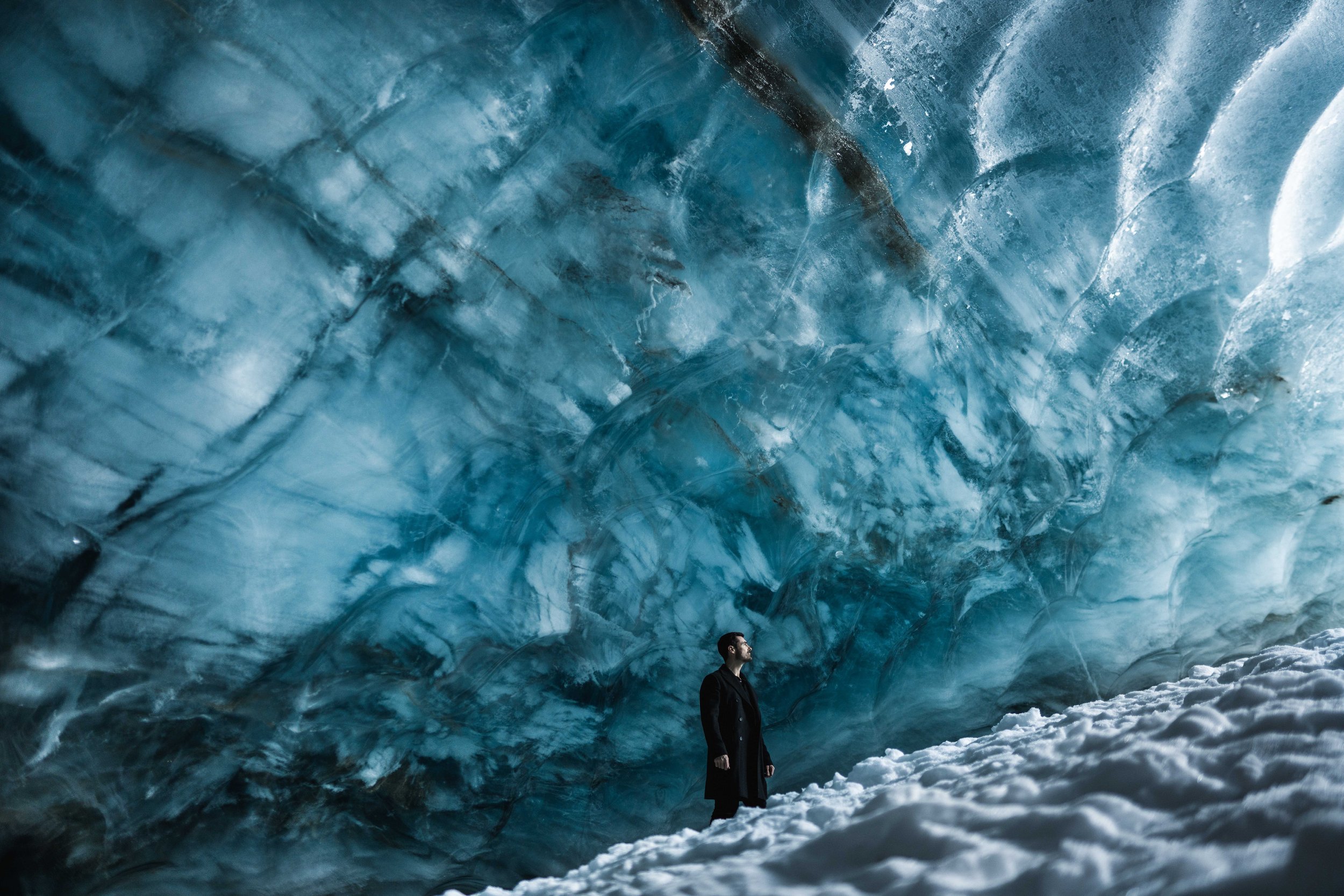A man in a black coat standing on snow inside an ice cave with blue-hued walls and ceiling.
