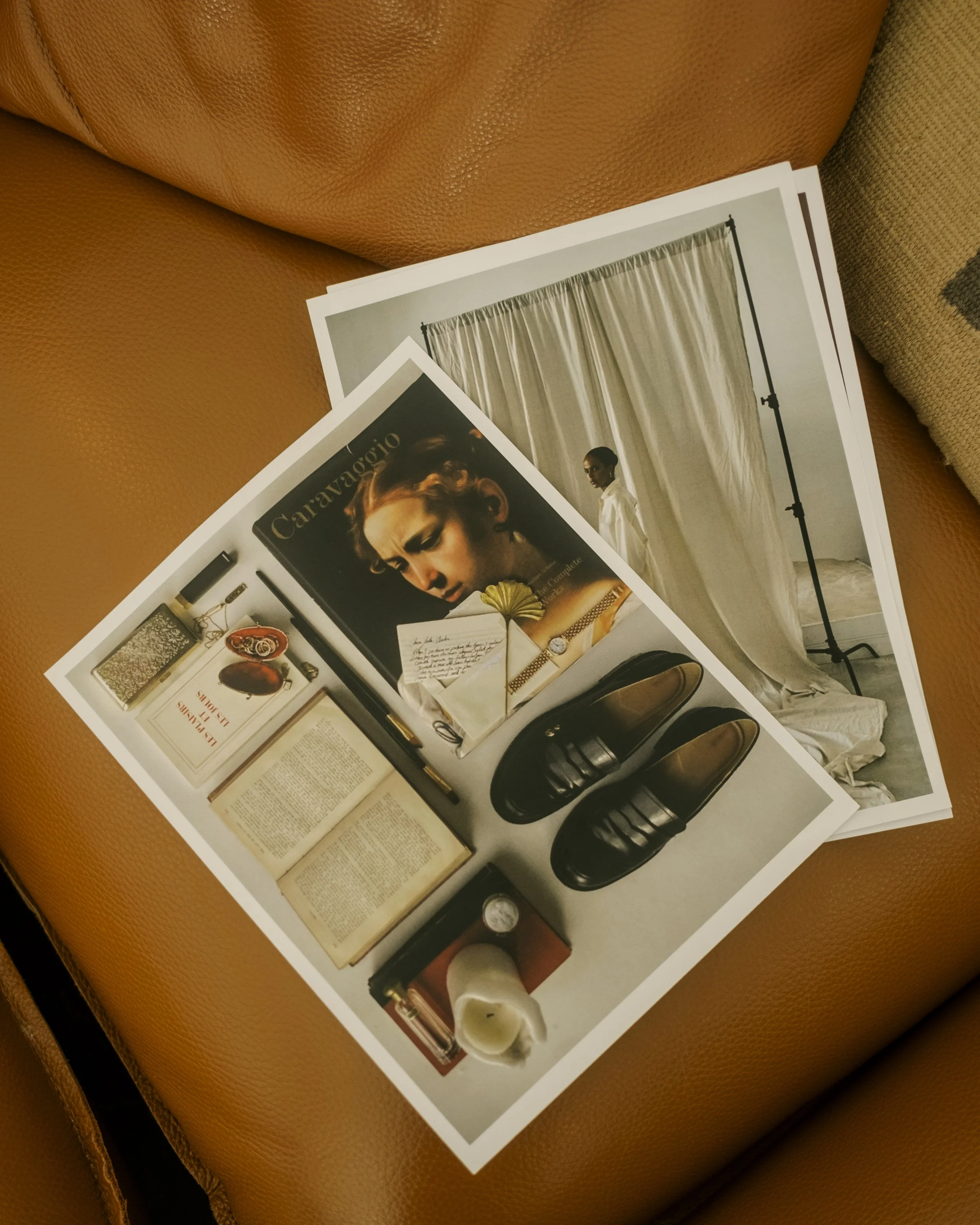 Photographs of various fashion and lifestyle items are laid out on a surface, including a Caravaggio art magazine, a book, shoes, a pen, an open book, a candle, and decorative objects, with a backdrop of a model in front of a curtain.