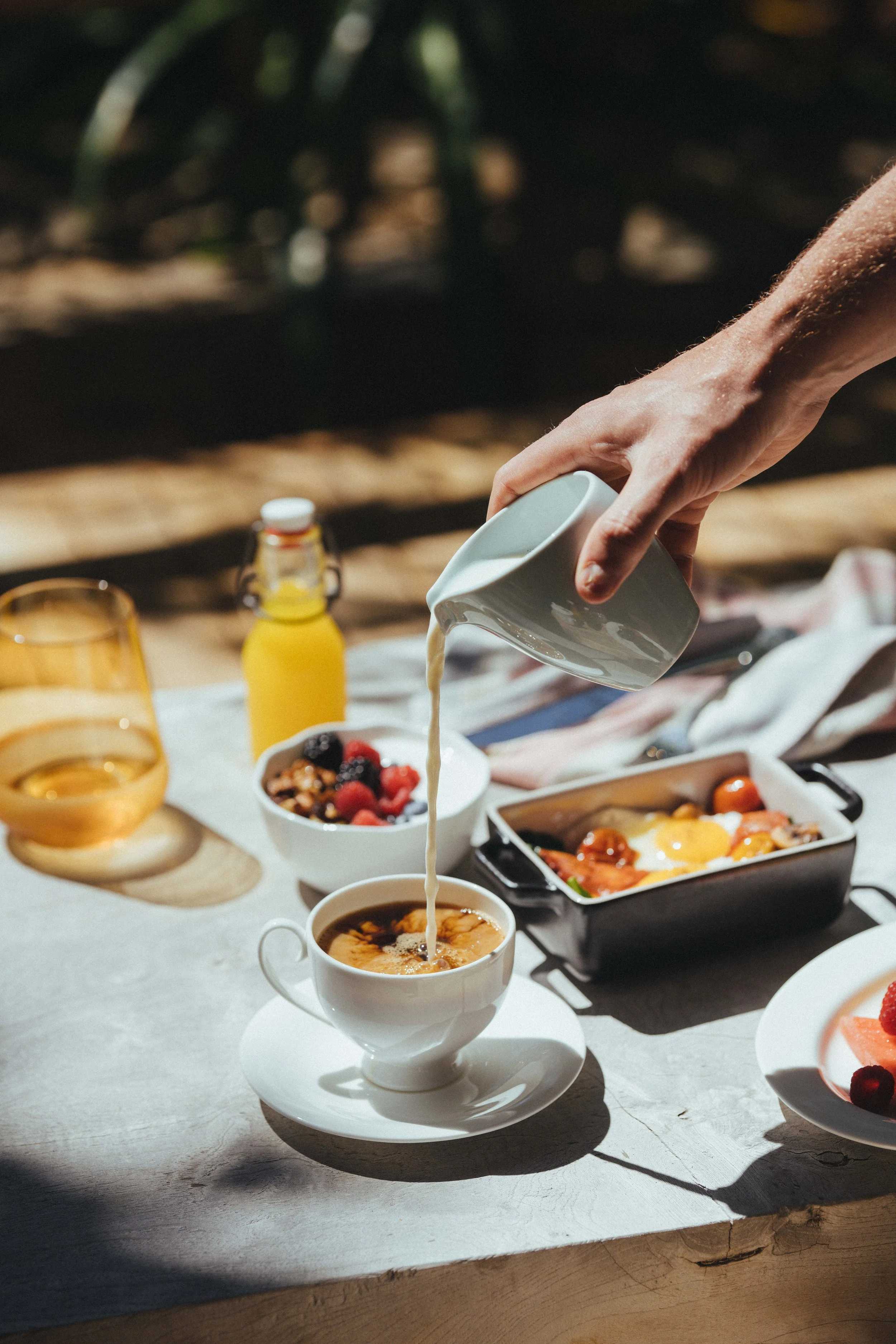 Person pouring cream into a cup of coffee on an outdoor table with breakfast foods.