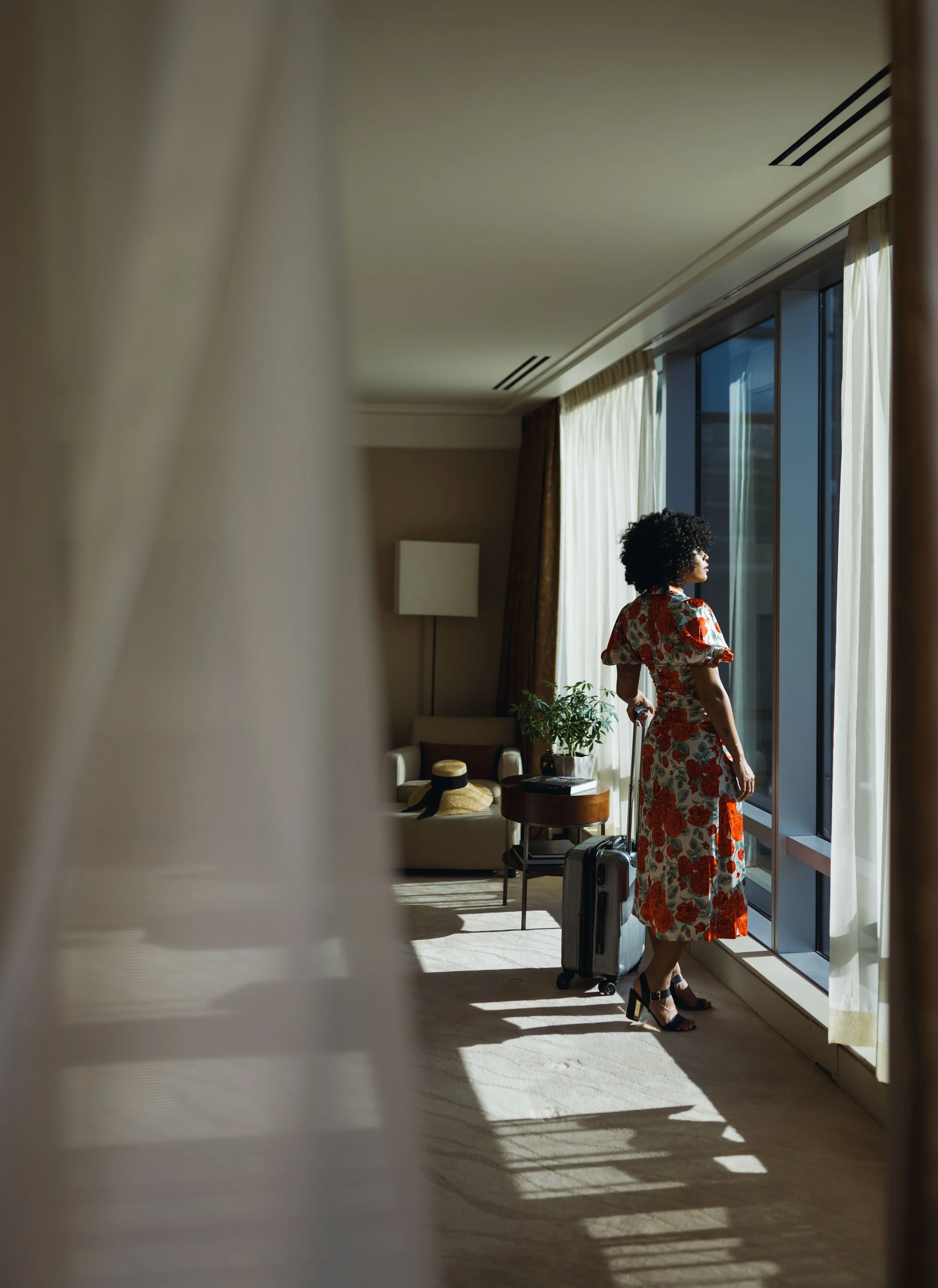 A woman with curly hair in a floral dress stands by a large window with sunlight streaming in, holding a suitcase in a hotel room.