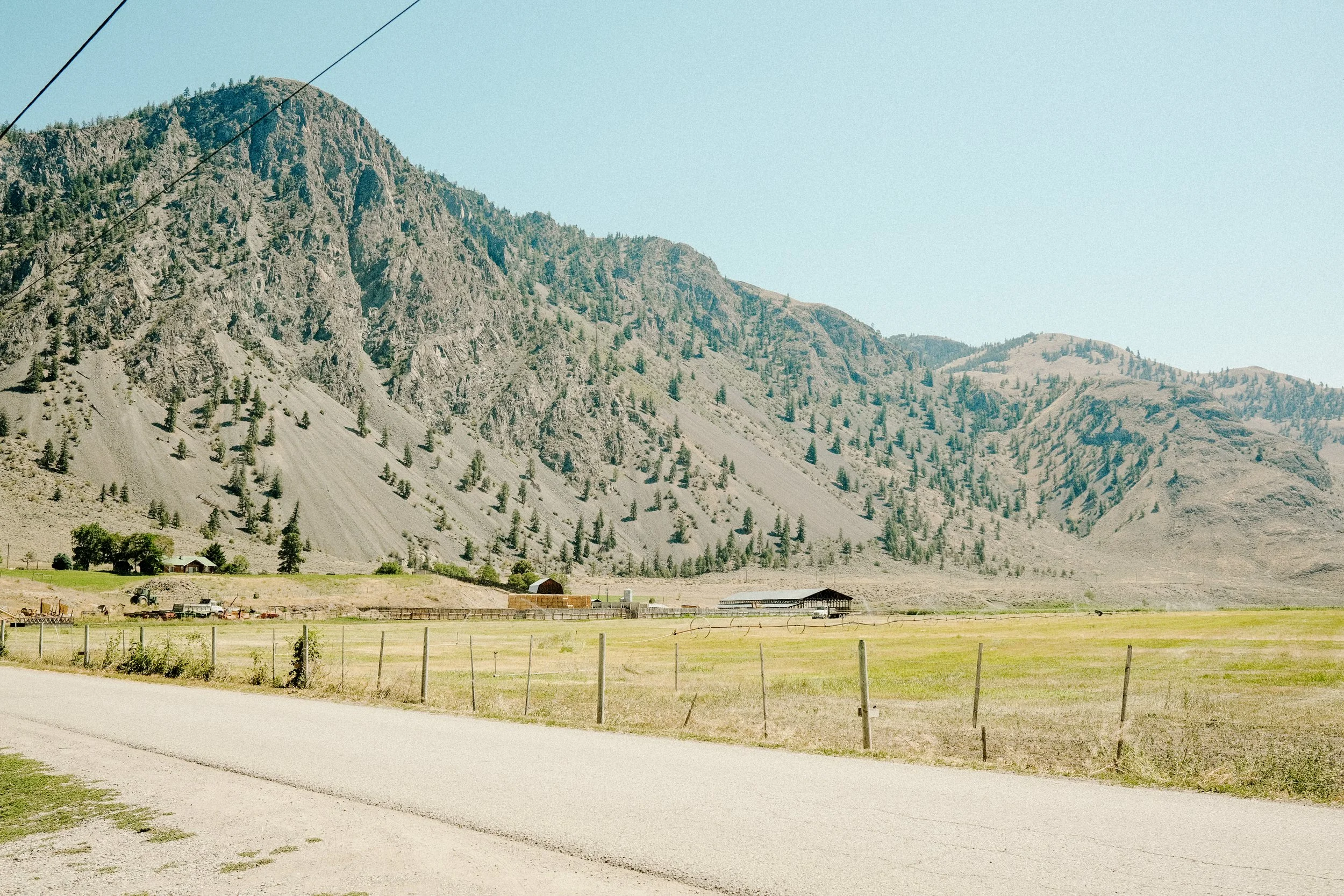 A rural landscape with a mountain range in the background, an open field with grass, and a dirt road in the foreground. There are fences and farm structures, including barns and machinery.