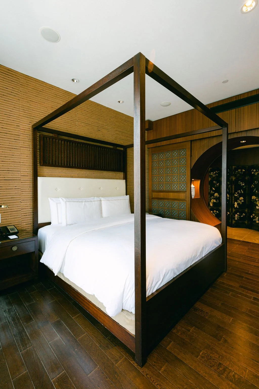 A hotel room with a four-poster bed, white bedding, wooden flooring, and Asian-inspired decor including wooden panels and a curved wooden screen.