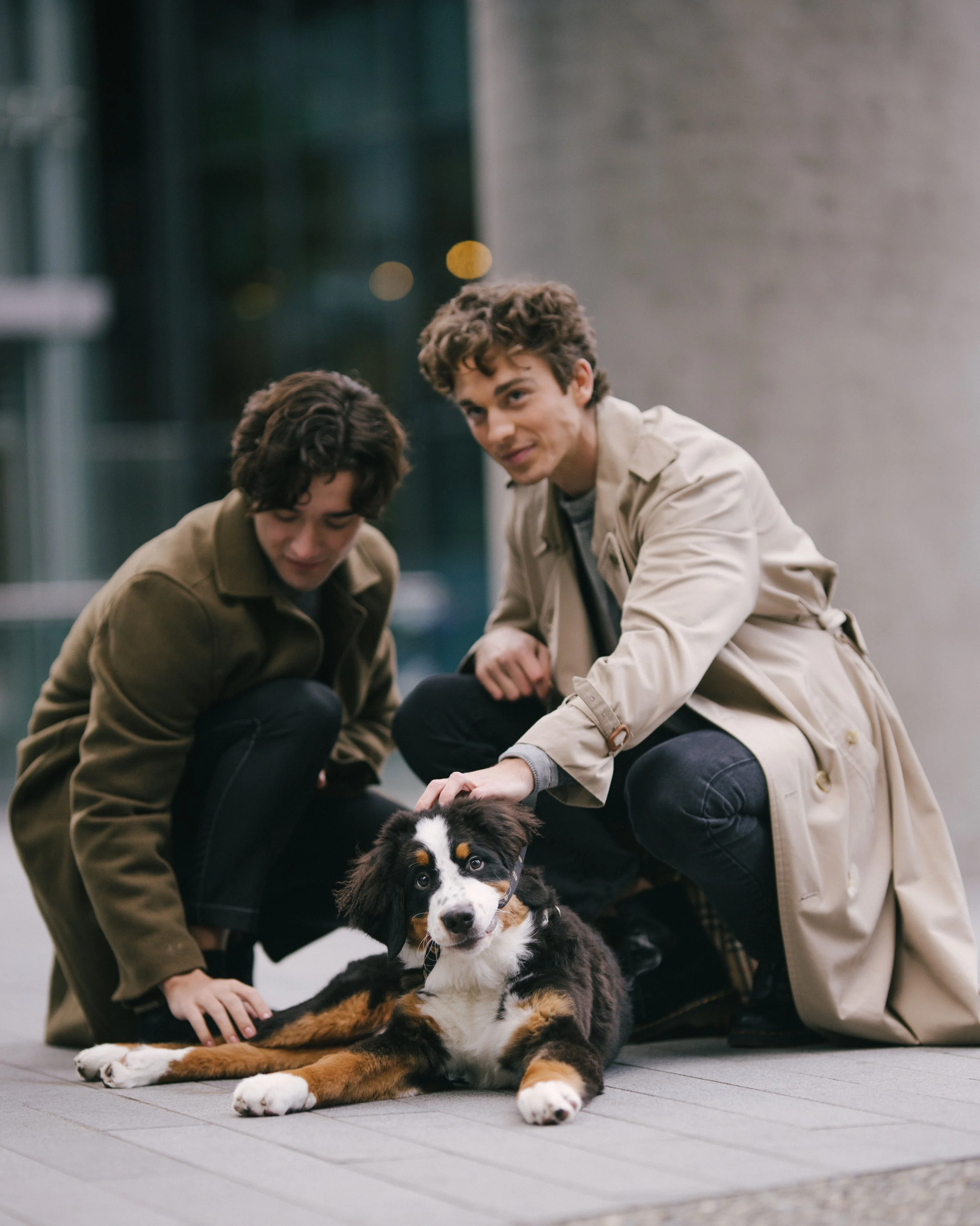 Two young men kneel beside a sitting Bernese Mountain Dog in an urban setting at night, with blurred city lights in the background.