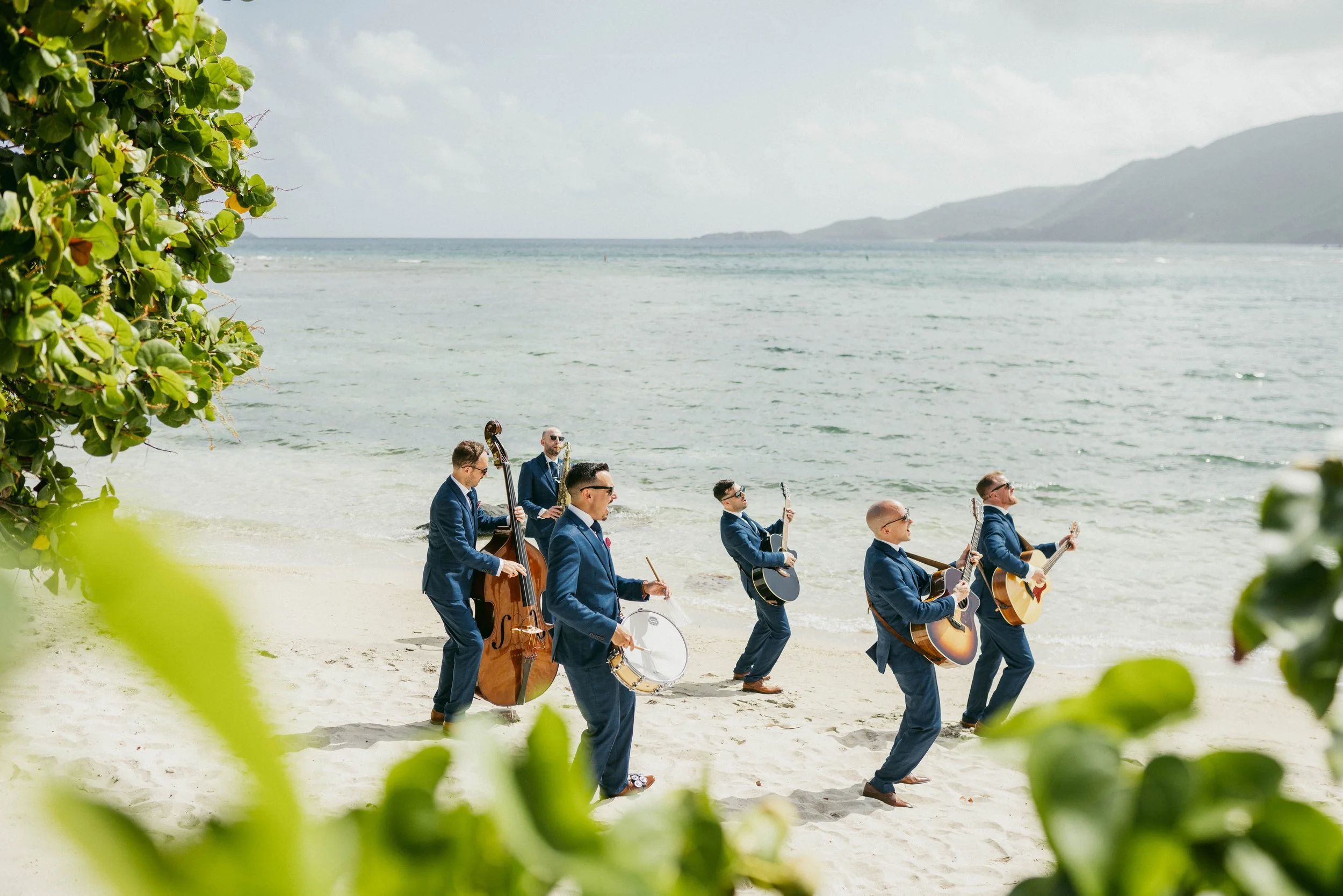 A band of seven men in blue suits playing instruments on the beach near the water, with mountains in the background and green foliage framing the scene.