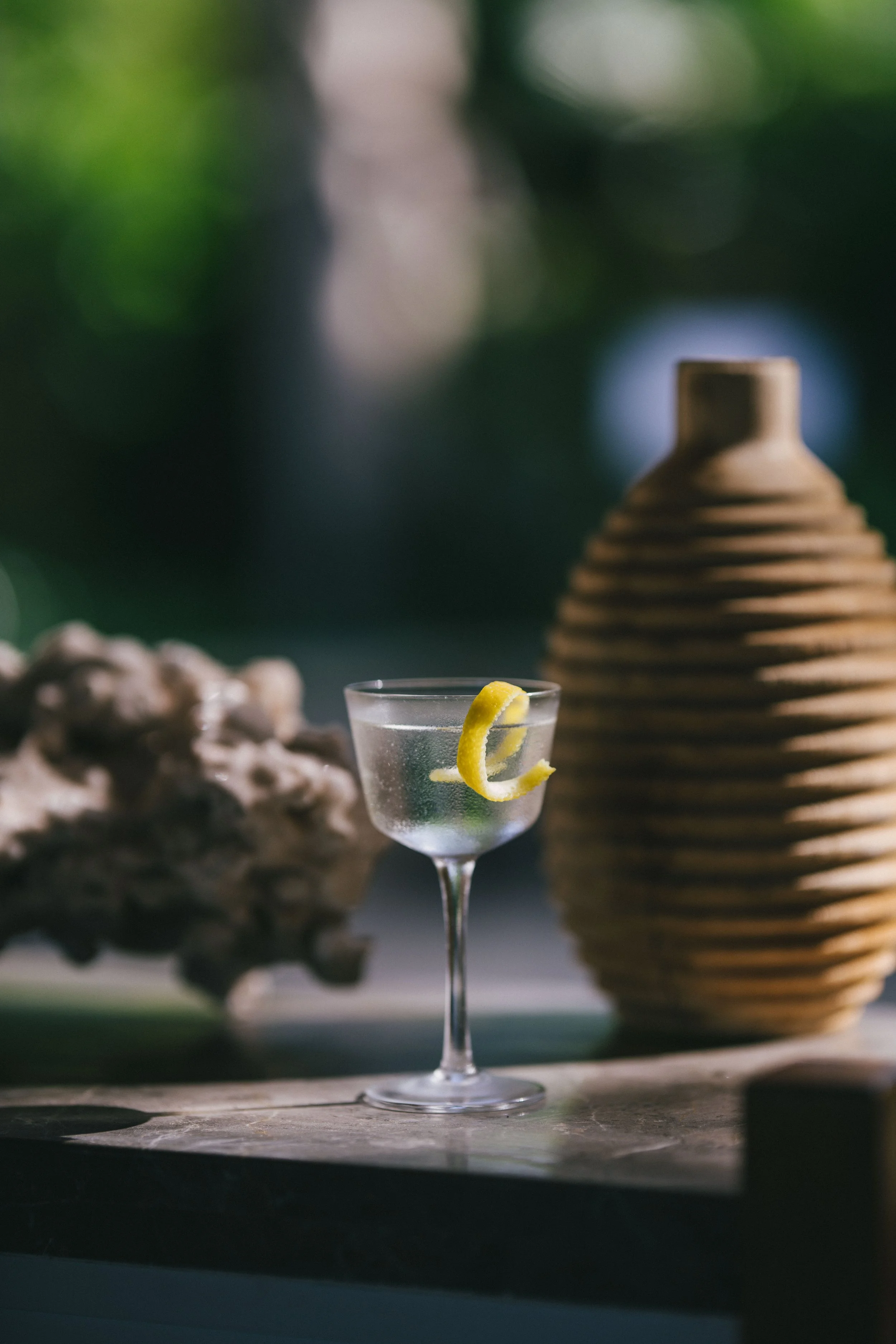 A cocktail glass with a lemon twist garnish on a wooden table, with a textured vase and natural background.