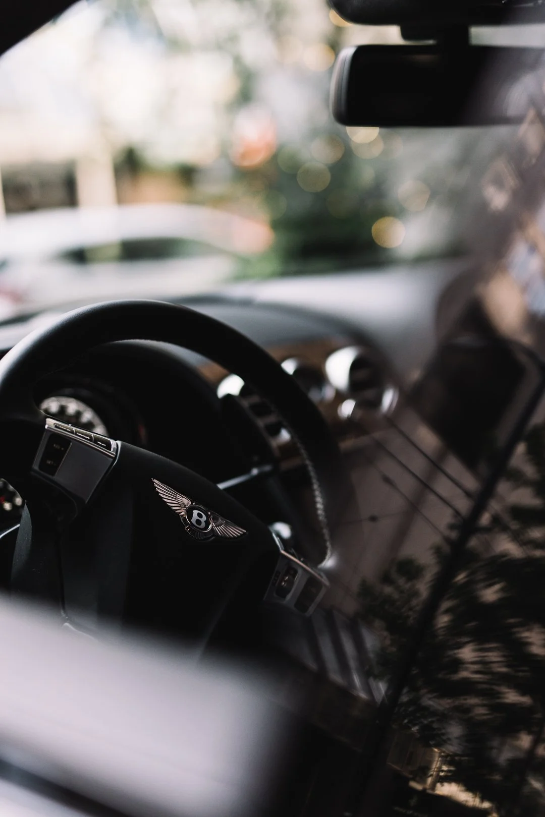 Close-up of a black Bentley steering wheel inside a car, with a blurred background of trees and lights.