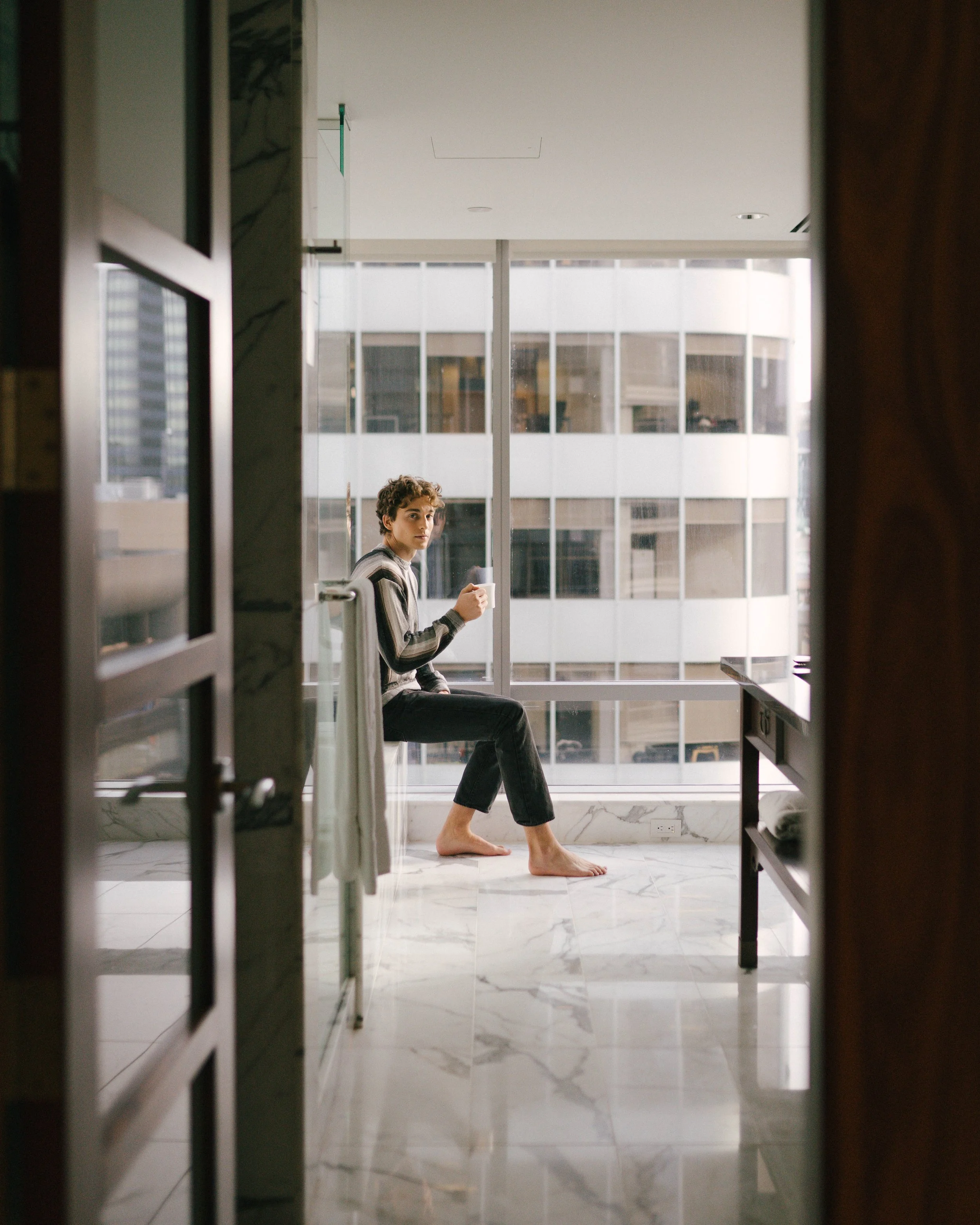 A person with curly hair, wearing a striped shirt and black pants, sitting barefoot on a windowsill and holding a white mug in a bright room with large windows, marble floor, and modern city buildings outside.