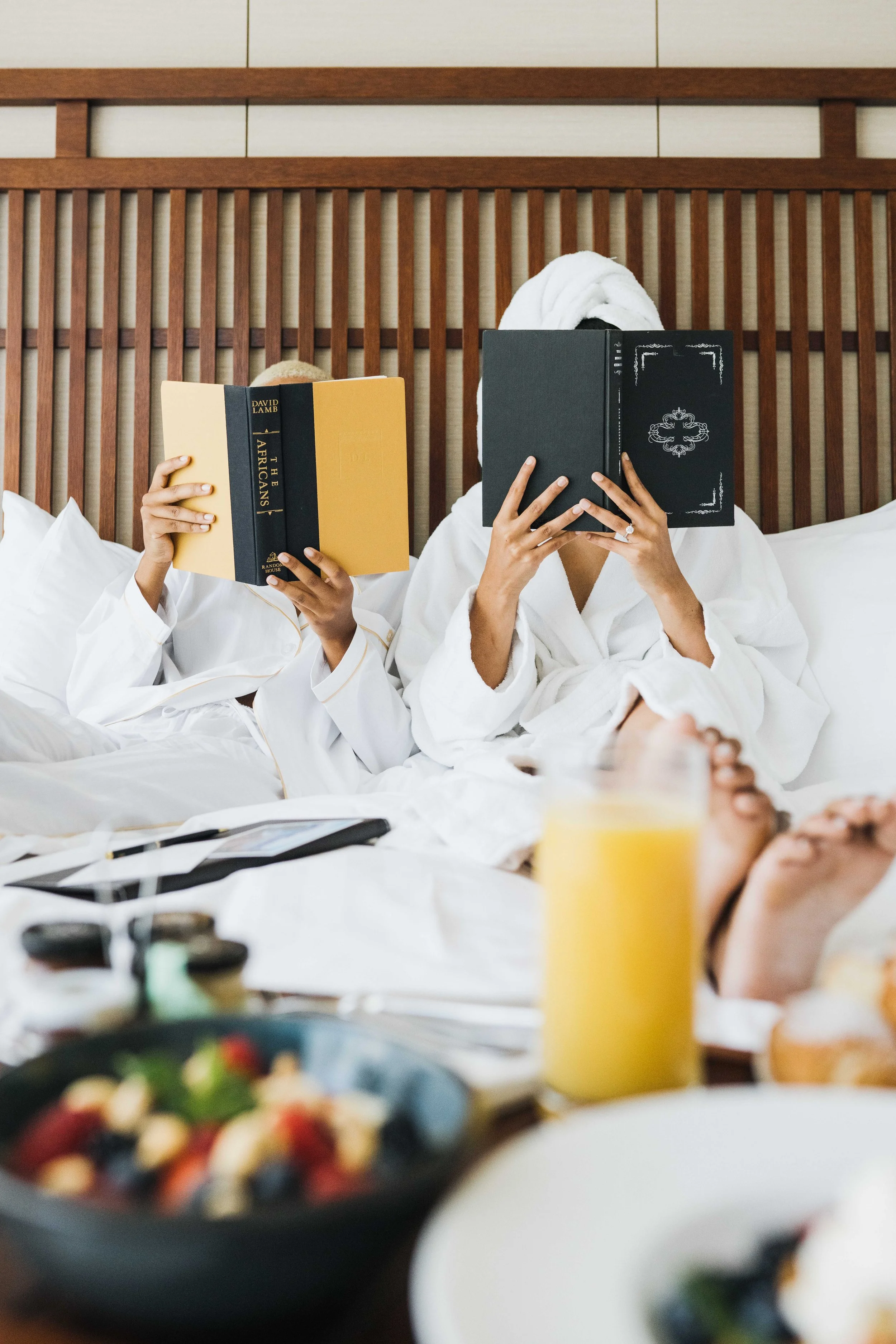 Two people in white robes lying on a bed, each reading a book, with a breakfast spread in front including a bowl of fruit and a glass of orange juice, and a wooden headboard behind them.