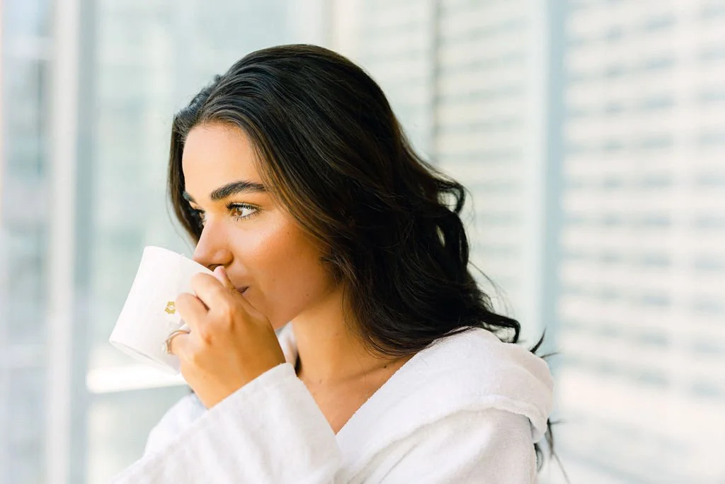 A woman with dark, wavy hair drinks from a white mug in a bright room with large windows