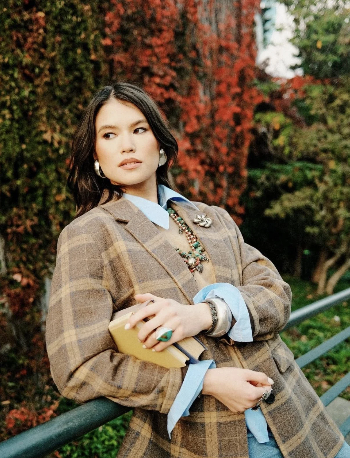 A woman with dark hair wearing a plaid blazer, layered necklace, large earrings, and rings, leaning on a green railing outdoors with red and green autumn foliage in the background.