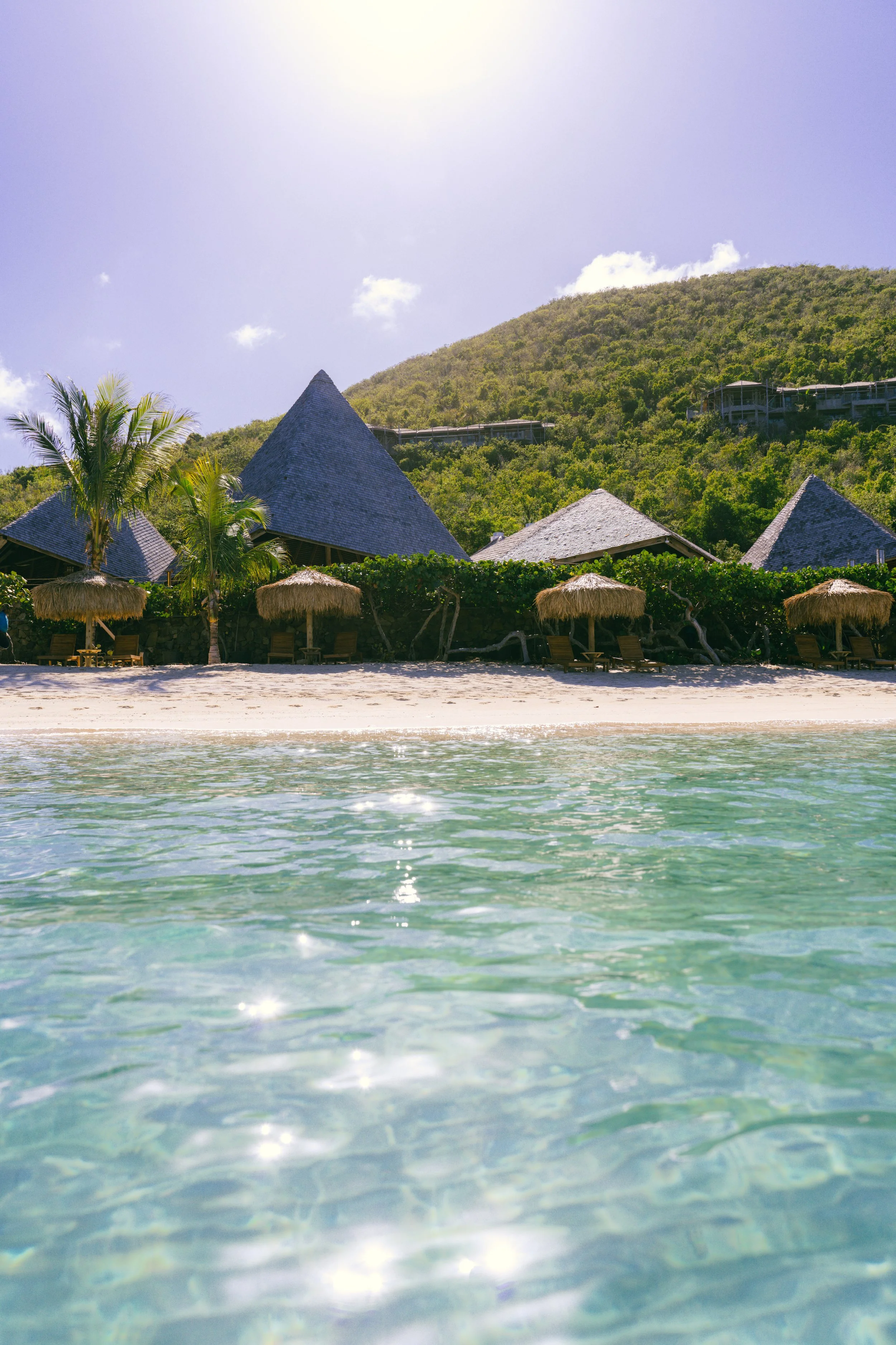 Beach with clear water, wooden lounge chairs, thatched umbrellas, palm trees, tropical huts, green hillside under a partly cloudy sky, bright sunlight.