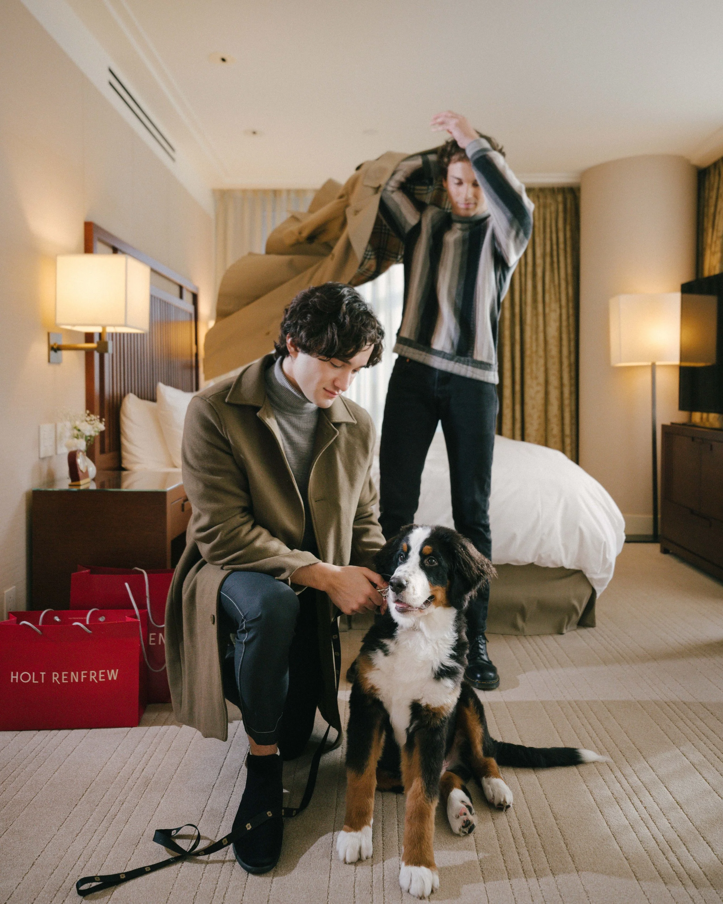 A young man is kneeling next to a happy Bernese mountain dog in a hotel room. Another person is in the background hanging up clothes on the bed's headboard. There are shopping bags on the floor with the text 'HOLT RENFREW'.