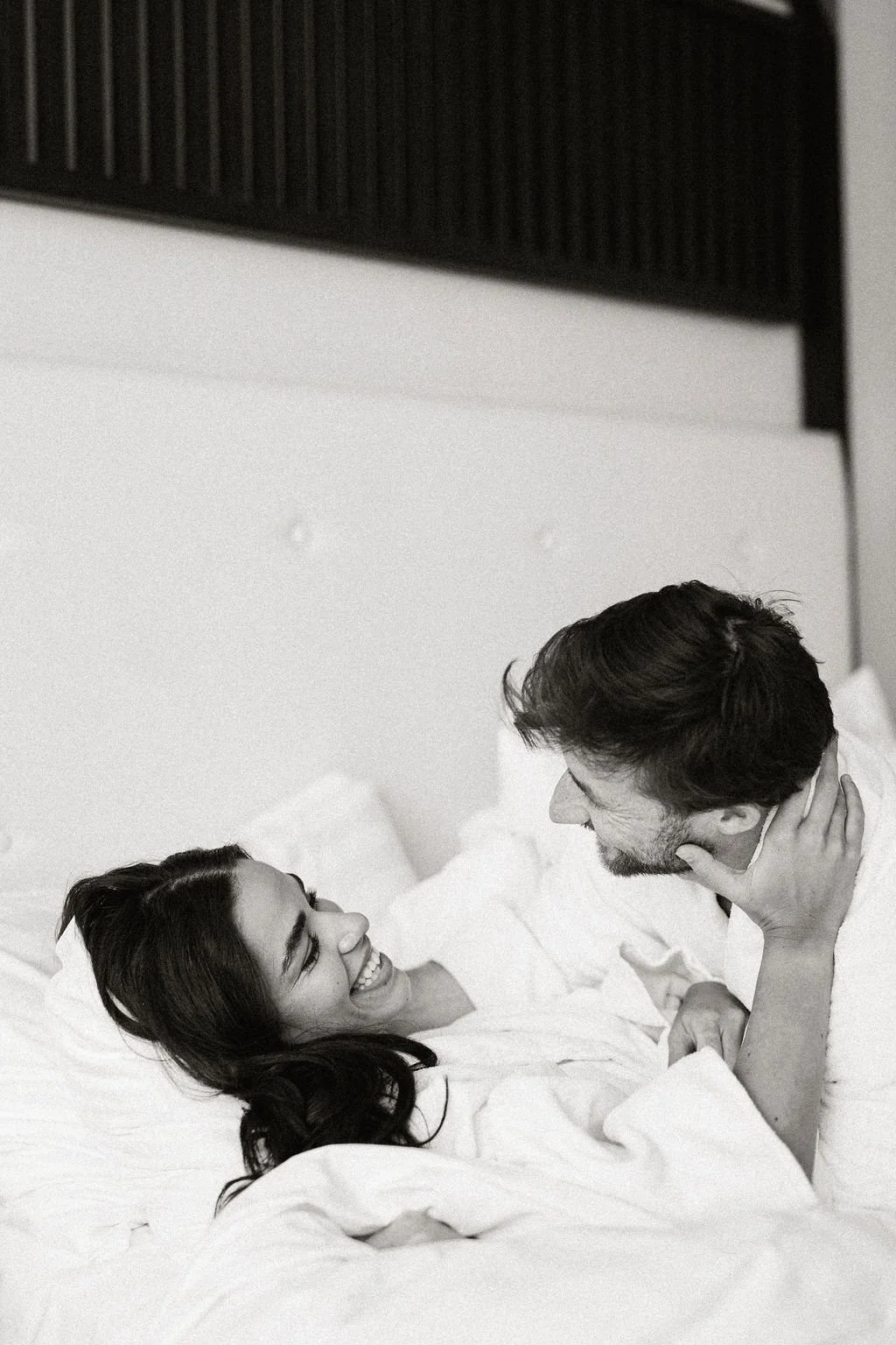 A woman and man lying in bed, smiling and looking at each other, with the woman touching the man's neck.