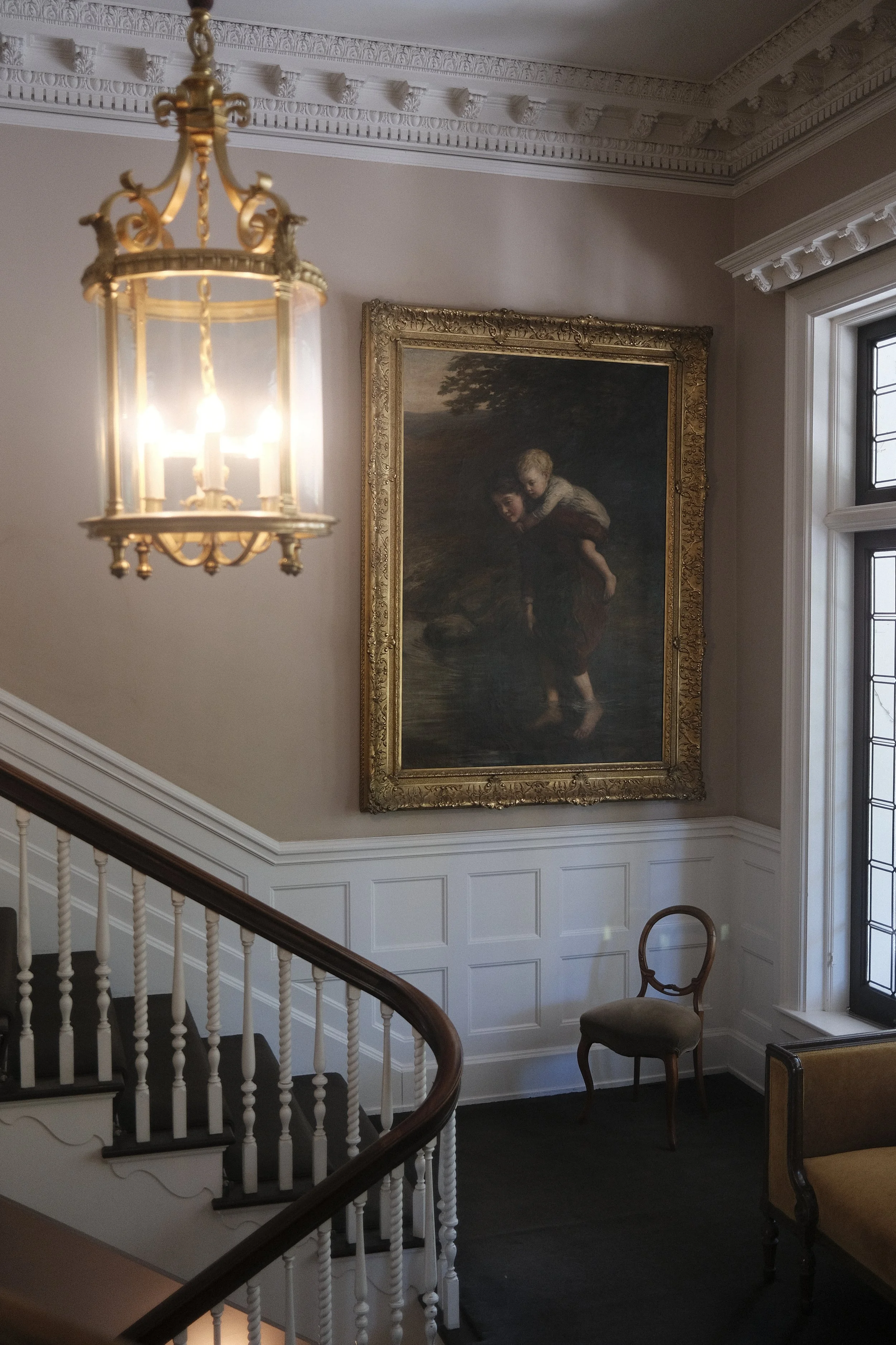 Interior of a historic room with an ornate chandelier, a large framed painting of children near water, a wooden staircase railing, and antique chairs near a large window.