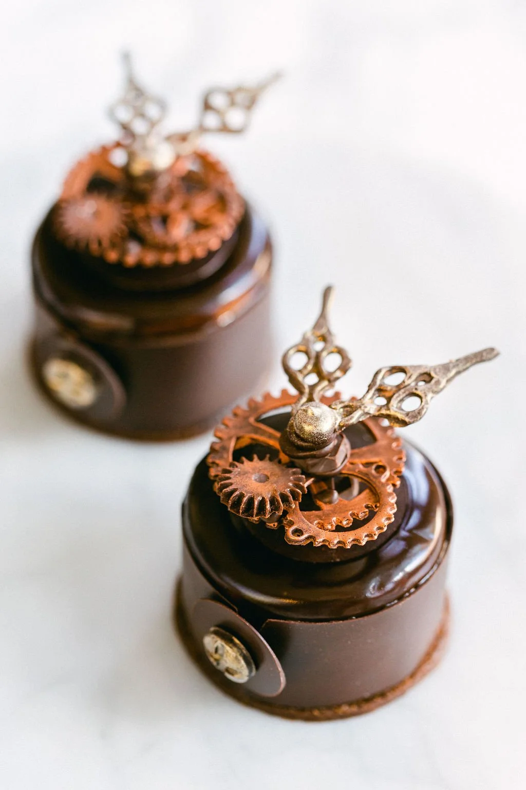 Two luxurious, round chocolate mousse desserts topped with chocolate gears and steampunk-style metallic decorations on a light background.