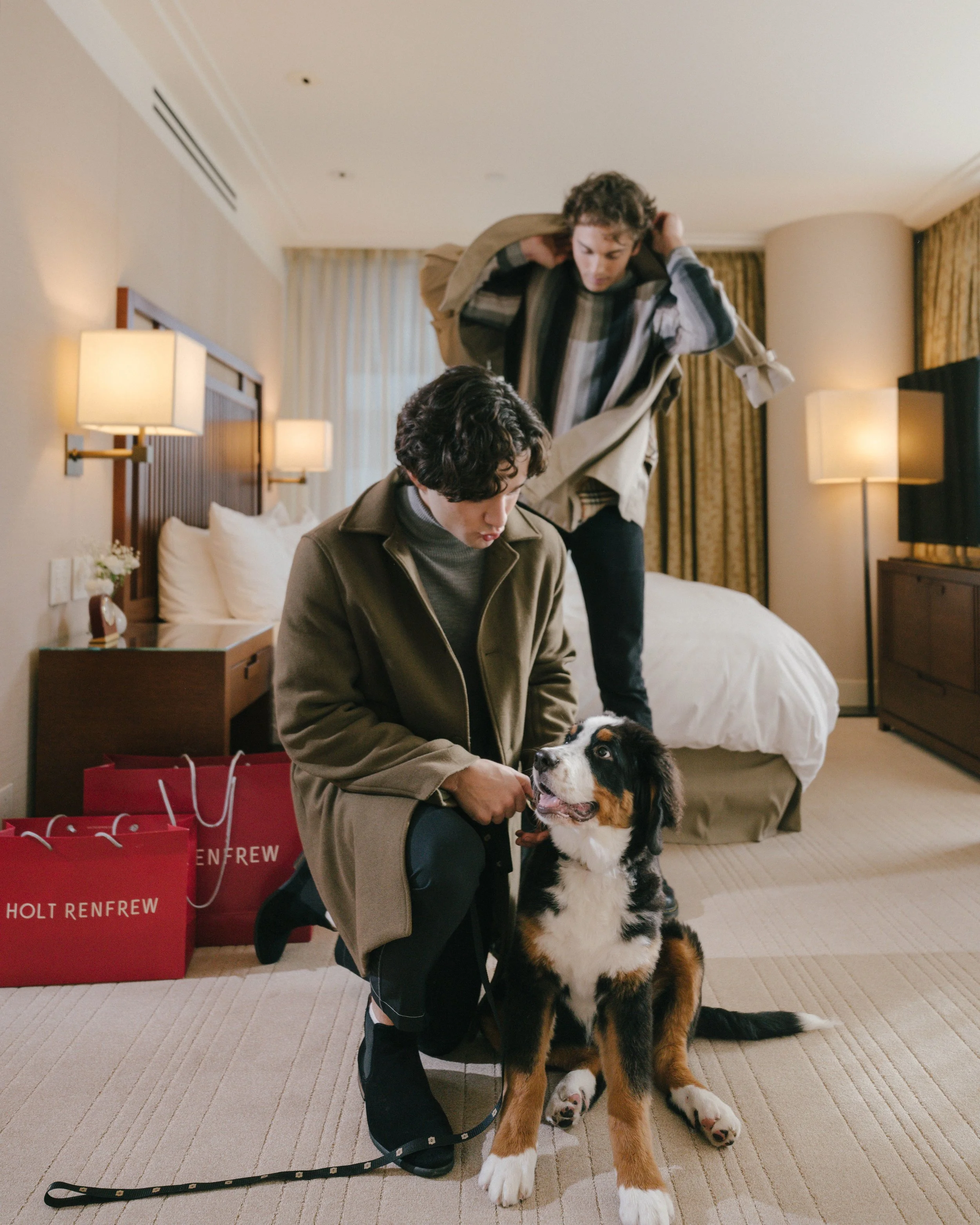 Two men and a dog inside a hotel room with beige carpet, bed, and lamps. One man is sitting on the floor with the dog, and the other is putting on a coat.