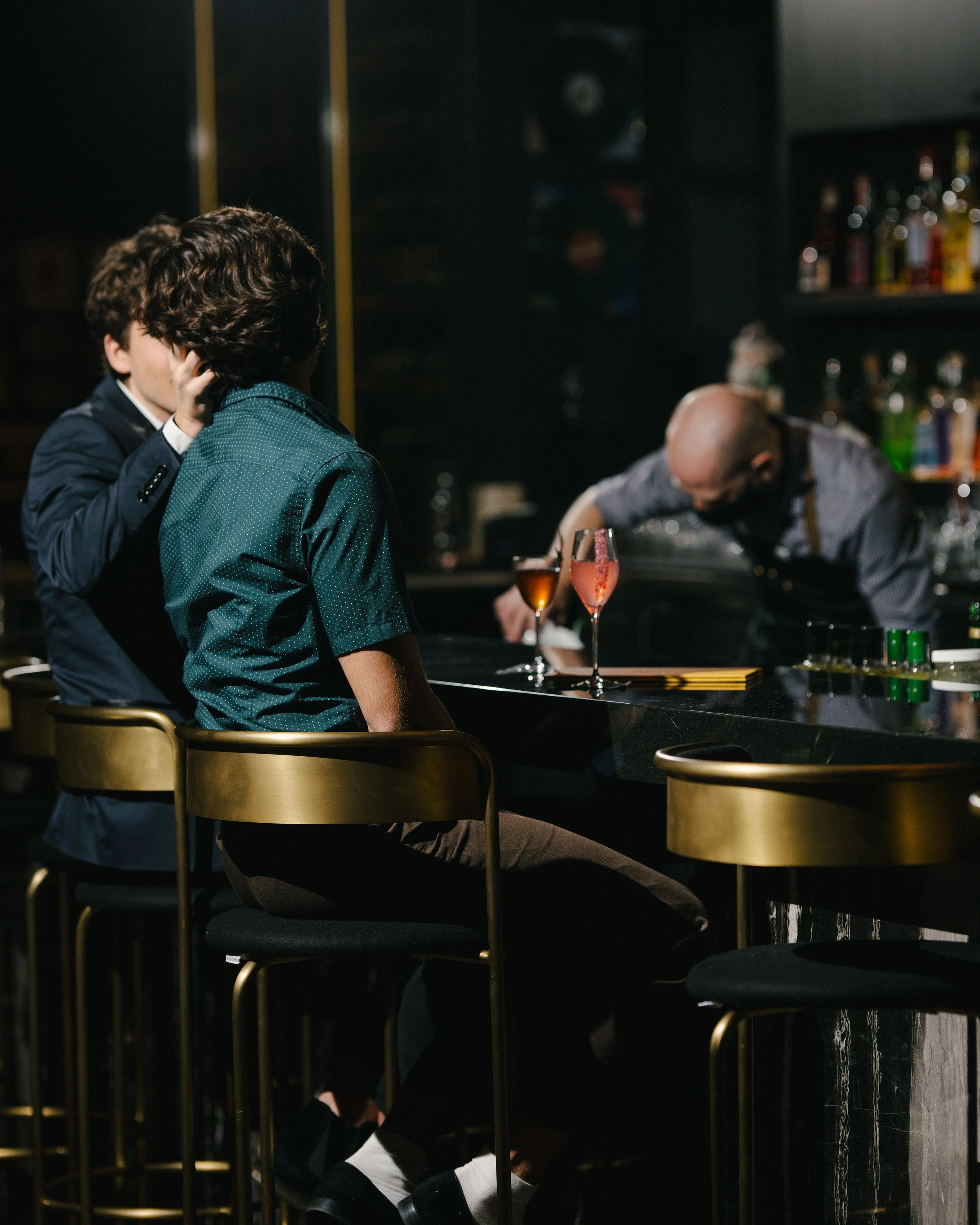 Two young men sitting at a bar, one in a blue suit, and the other in a teal shirt, with a bartender preparing drinks in the background, bar shelves with bottles, dim lighting.