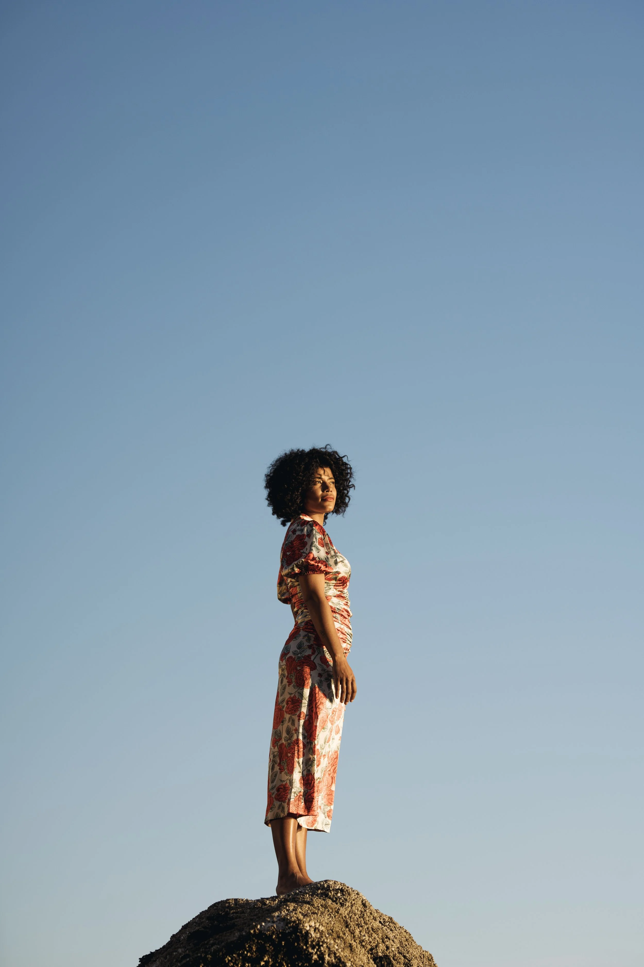 A woman in a floral dress standing on a rocky surface under a clear blue sky.