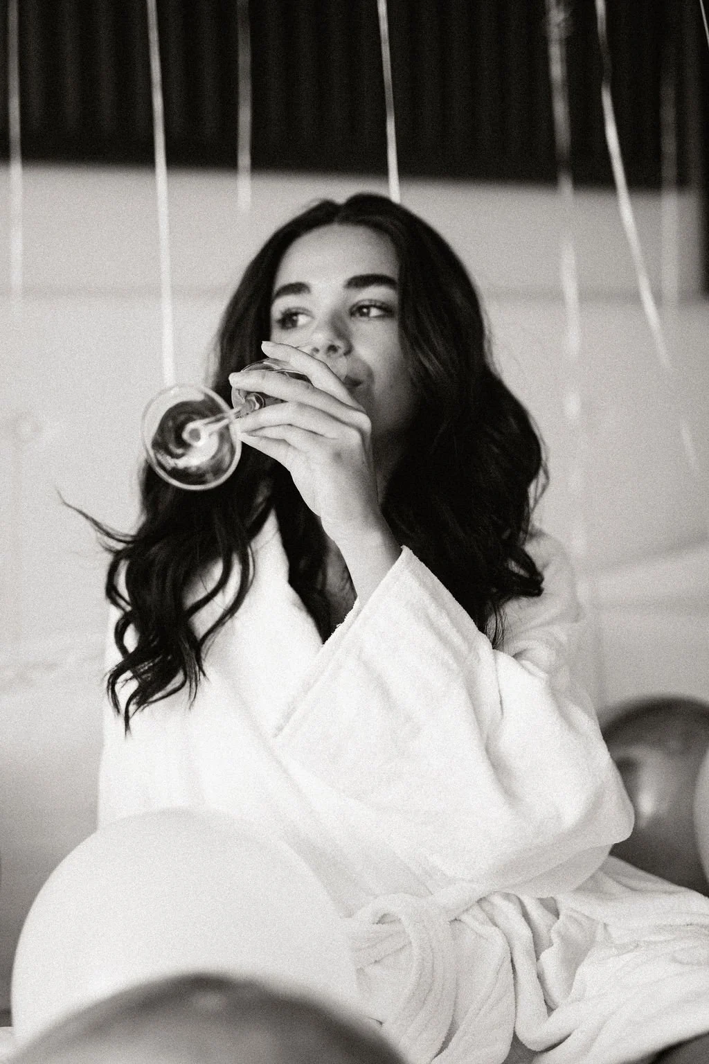 A black-and-white photo of a woman with long wavy hair sitting and drinking from a champagne glass, wearing a white bathrobe.