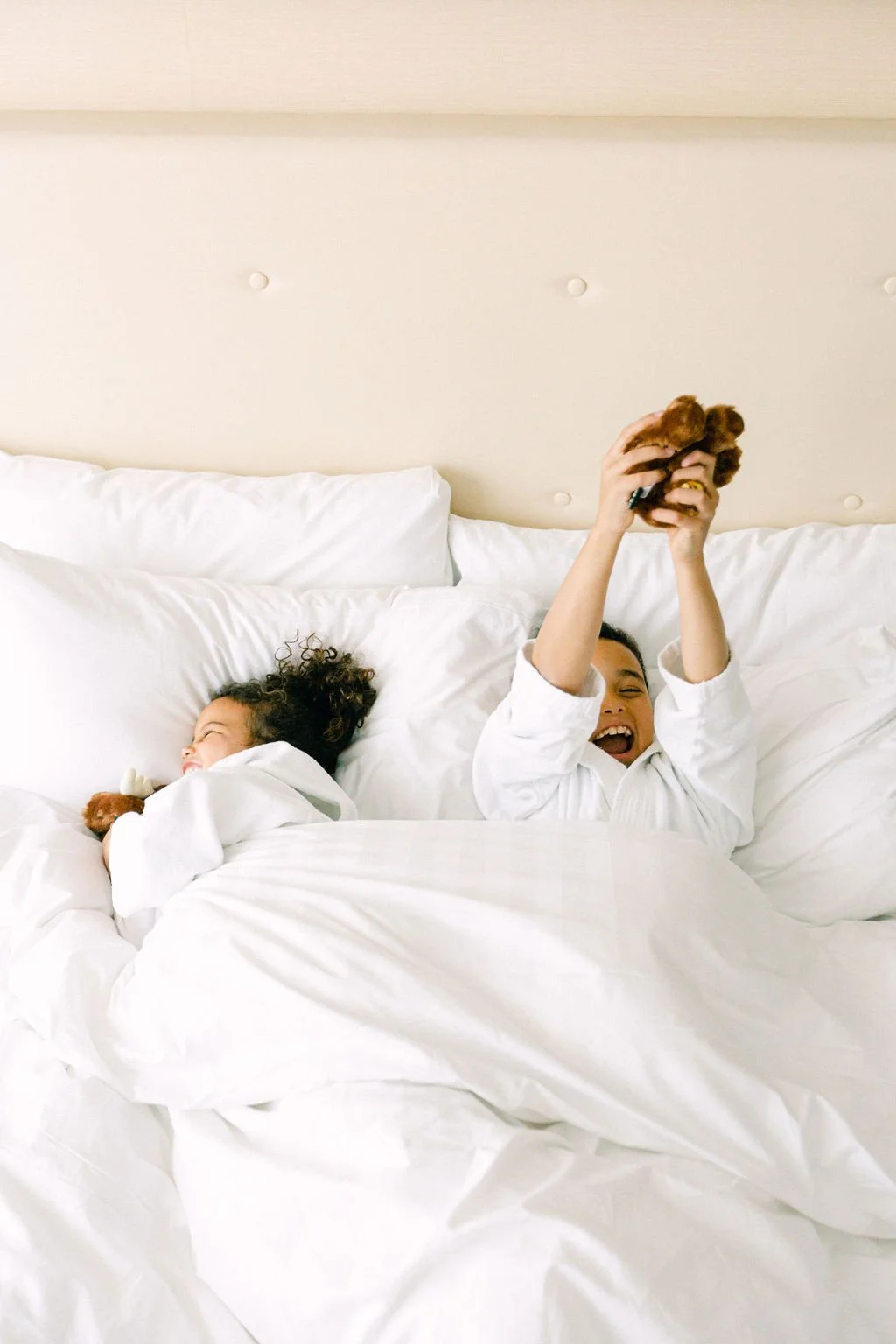 Two children, a girl and a boy, are lying in bed with white sheets and pillows, playing with stuffed animals. The girl is sleeping, and the boy is laughing while holding up a stuffed animal.