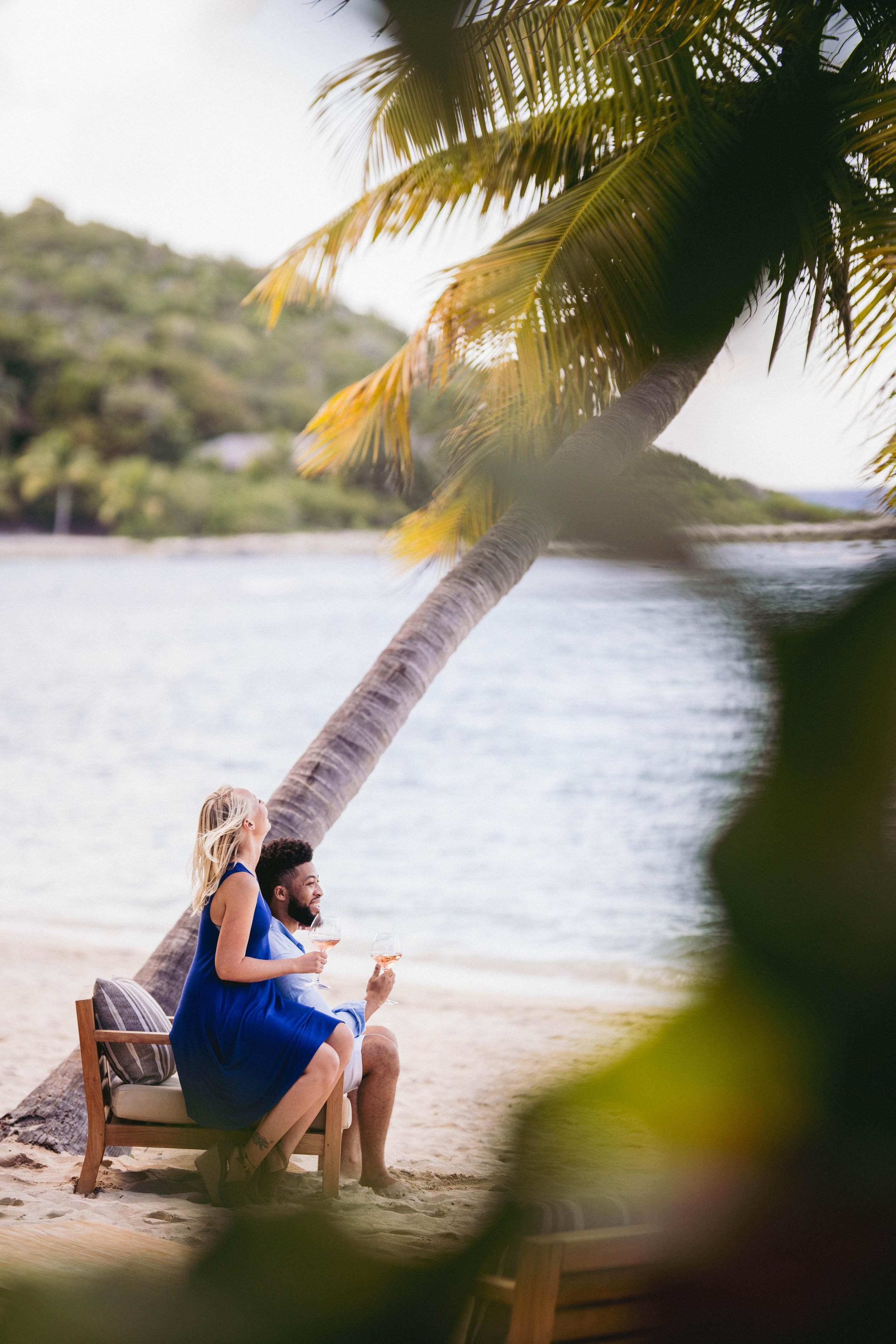 A woman and man sitting on a bench under a leaning palm tree on a beach, enjoying drinks, with water and hills in the background.