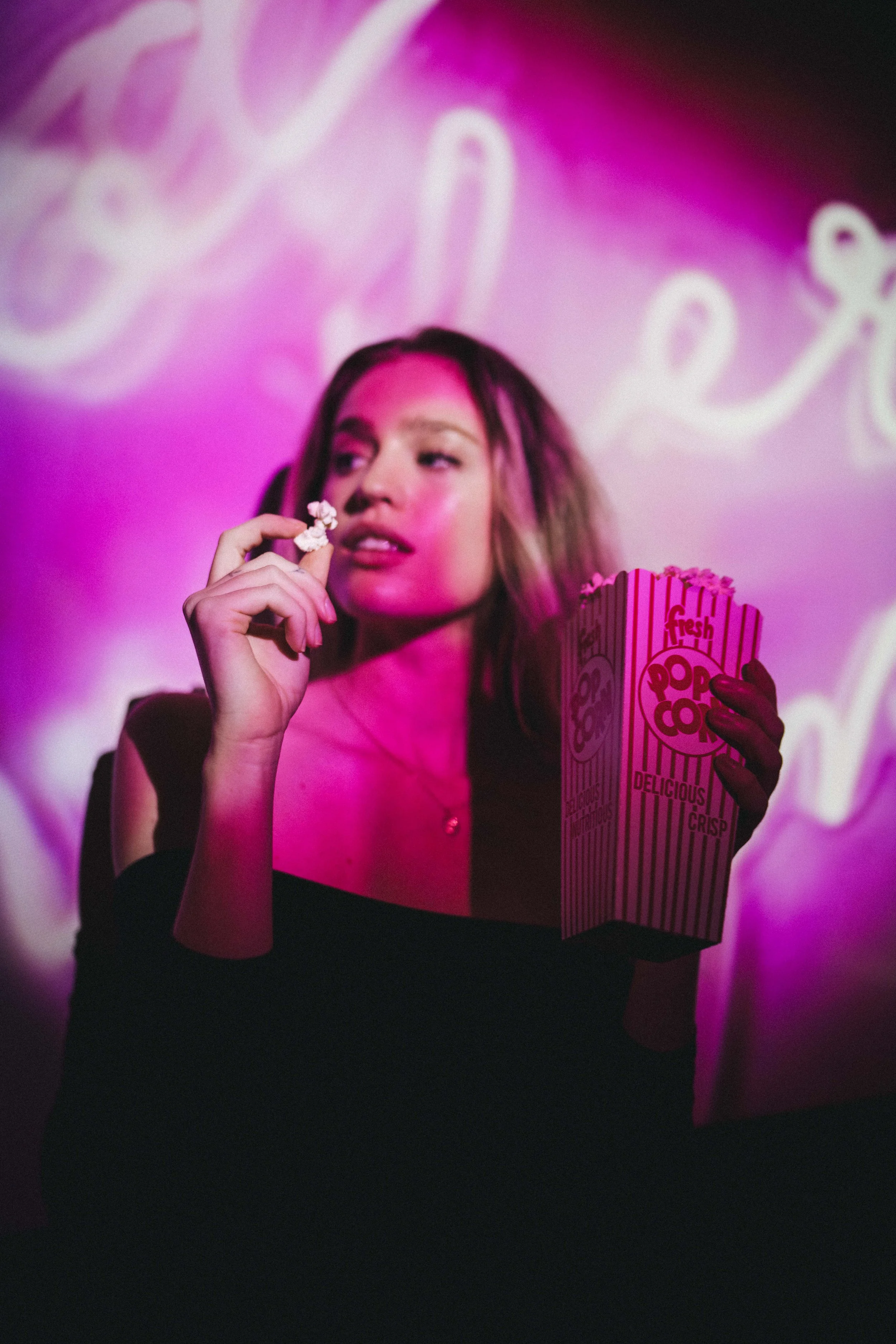 A woman with shoulder-length hair holding a box of popcorn in front of a pink-lit background with neon-style writing.