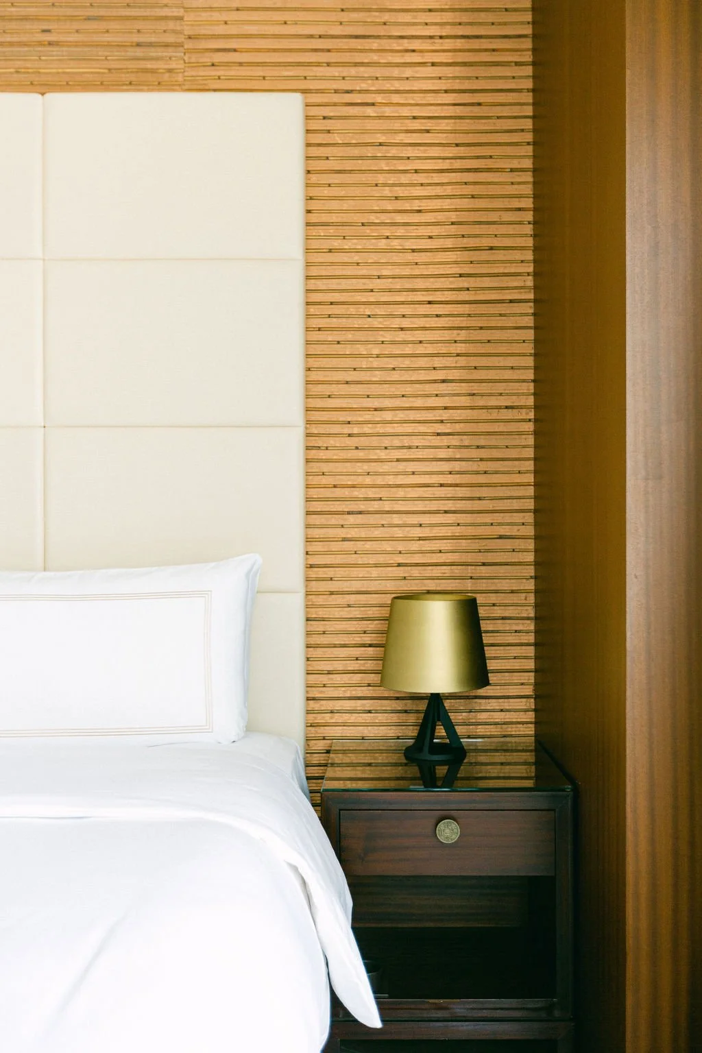 A bedroom corner with a white headboard, a white pillow, a dark wooden nightstand, and a gold and black table lamp against a wooden slat wall.