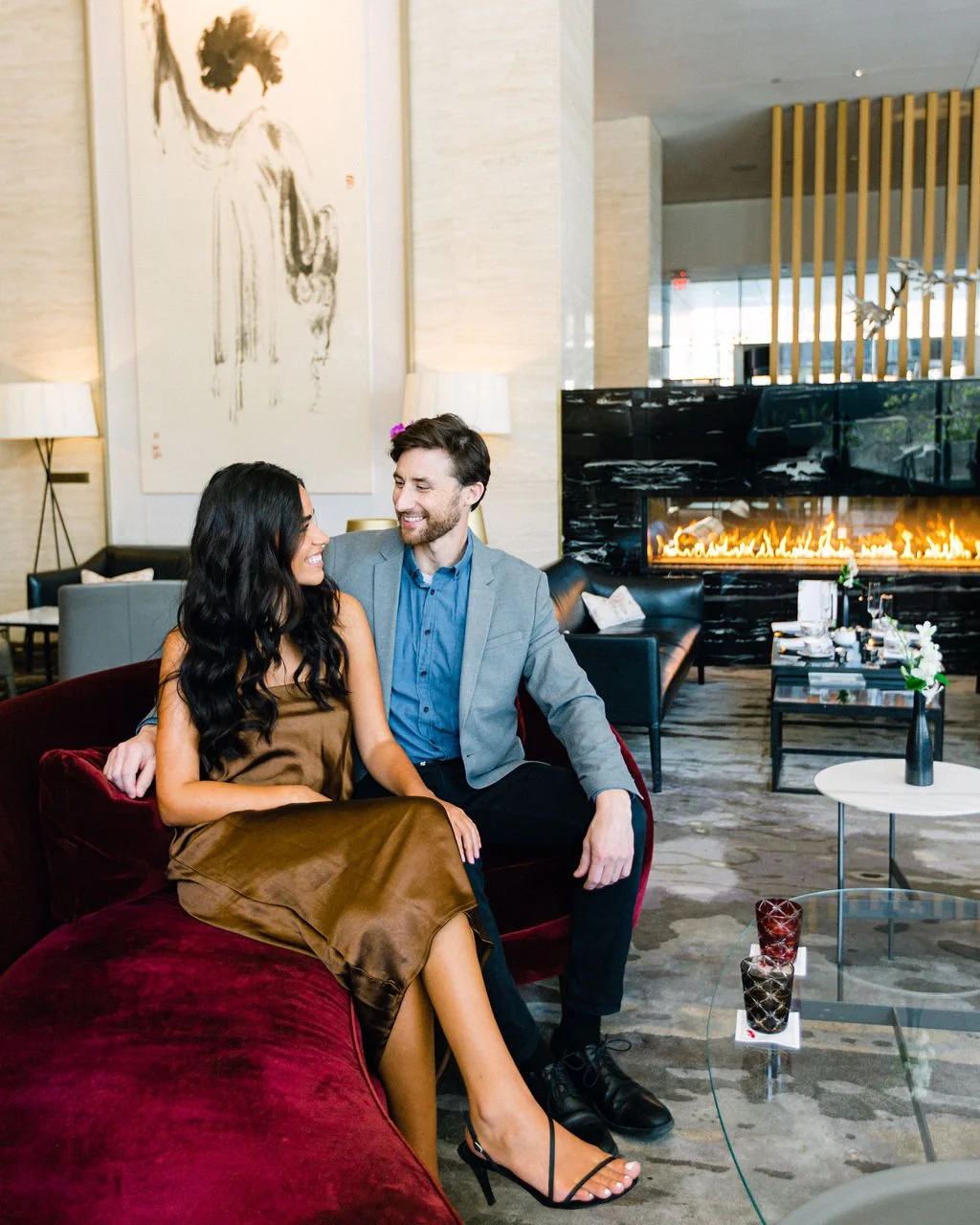 A smiling couple sitting close together on a velvet sofa in a modern hotel lobby with a fireplace behind them, elegant artwork on the wall, and contemporary decor.