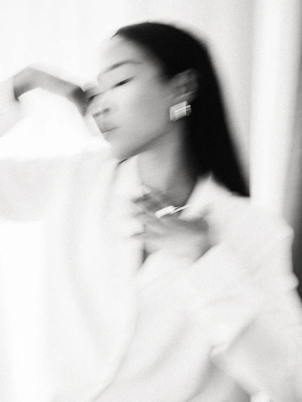 A blurred black-and-white photo of a woman with long dark hair, wearing earrings, resting her head on her arm.