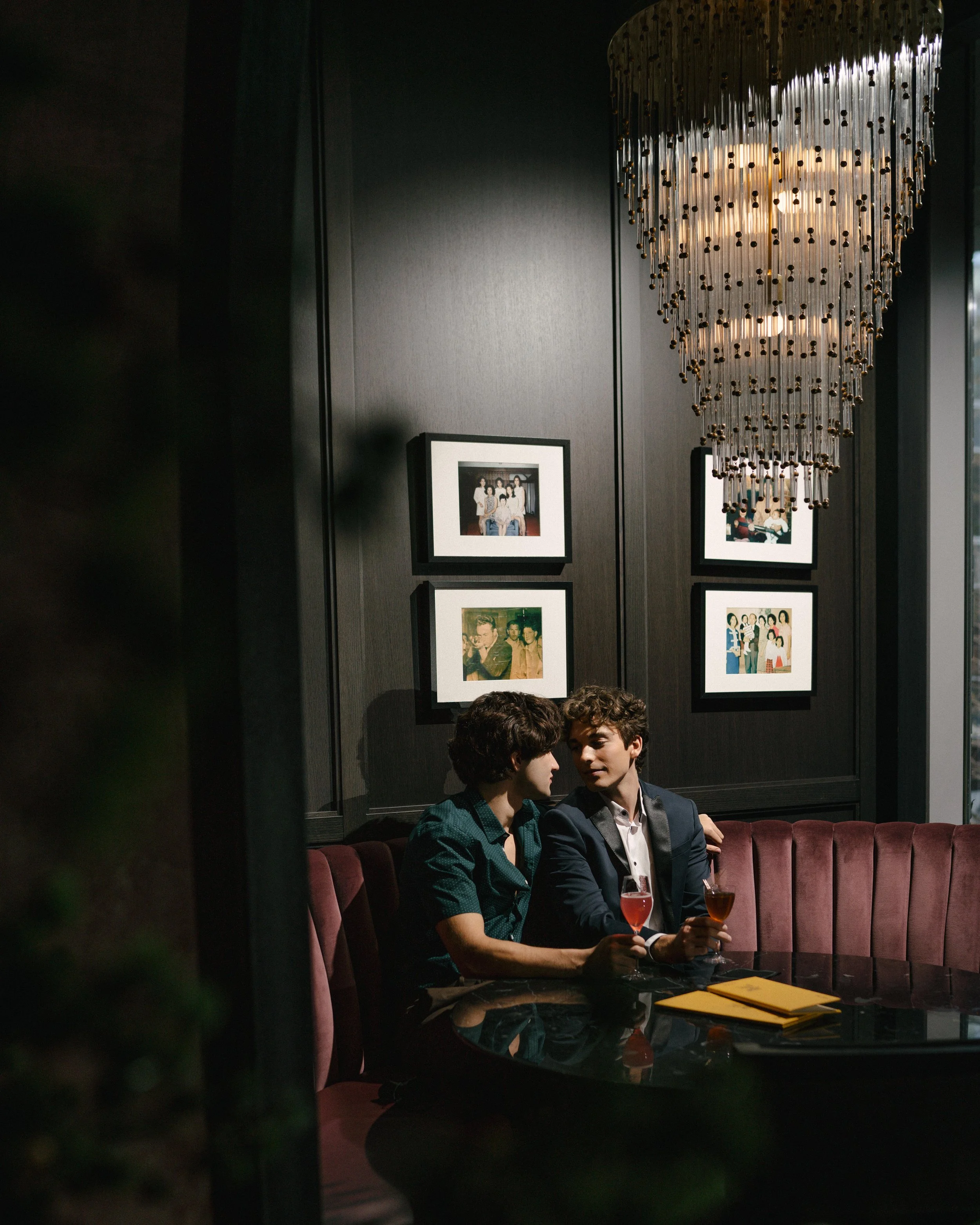 Two young men sitting at a round table in a dimly lit restaurant or lounge, holding glasses of drinks, with framed photographs on the dark wall behind them and a large chandelier hanging overhead.