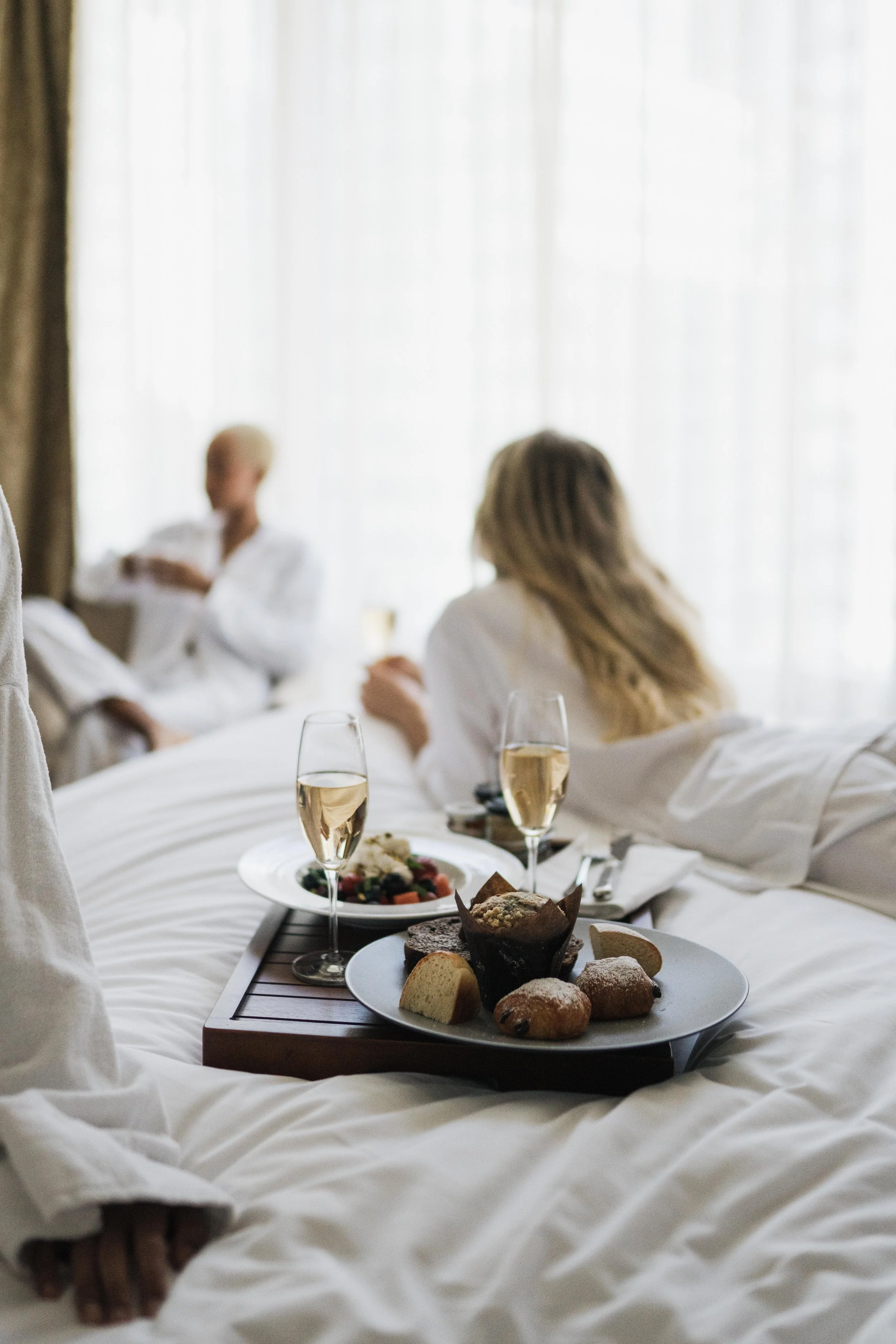 Tray with assorted desserts and two glasses of champagne on a bed, with three women in bathrobes in the background.