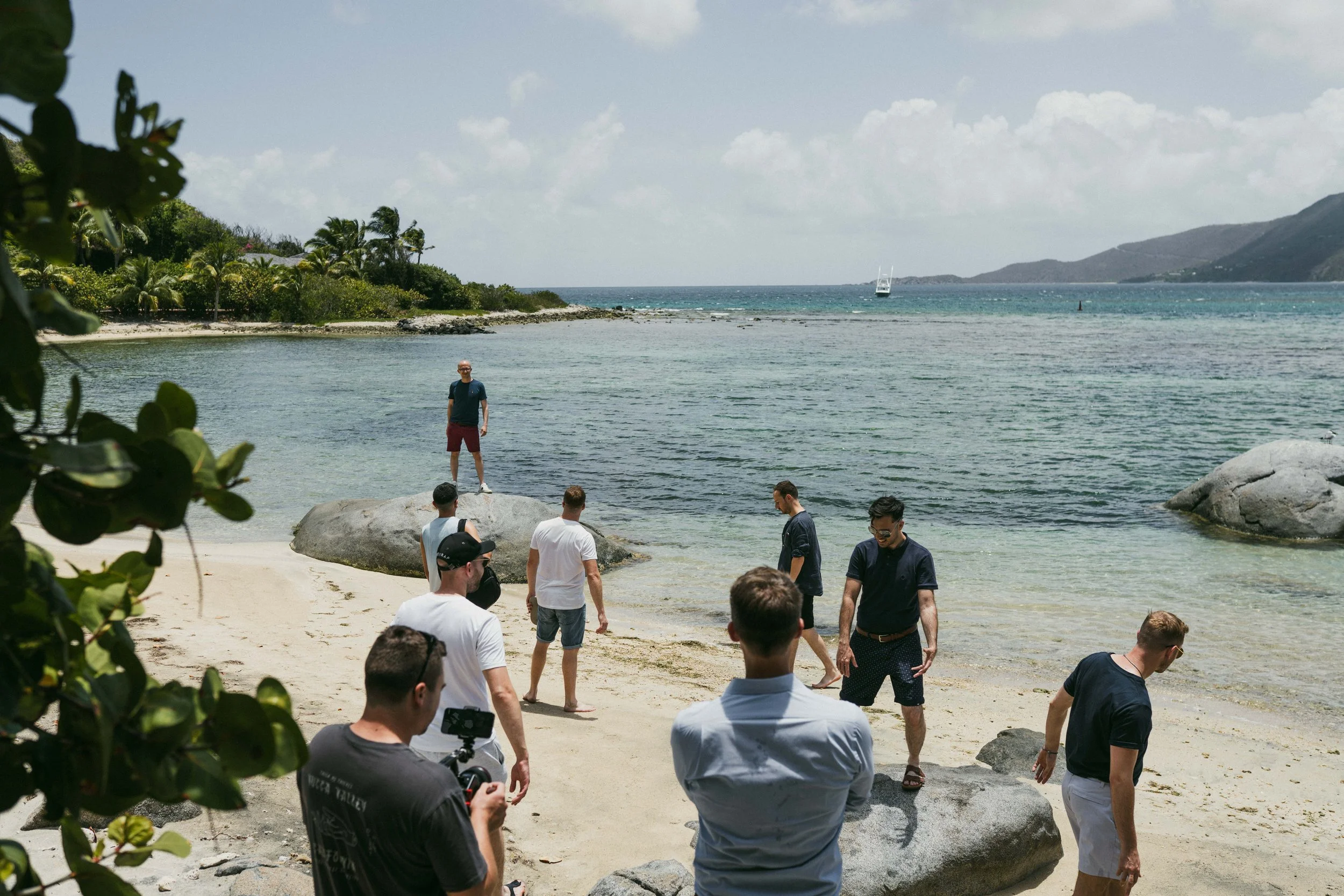 A group of people on a beach by the water, some standing on rocks, others walking or standing on the sand. There is a man standing on a large rock in the background, and a boat is visible on the water. The sky is partly cloudy.