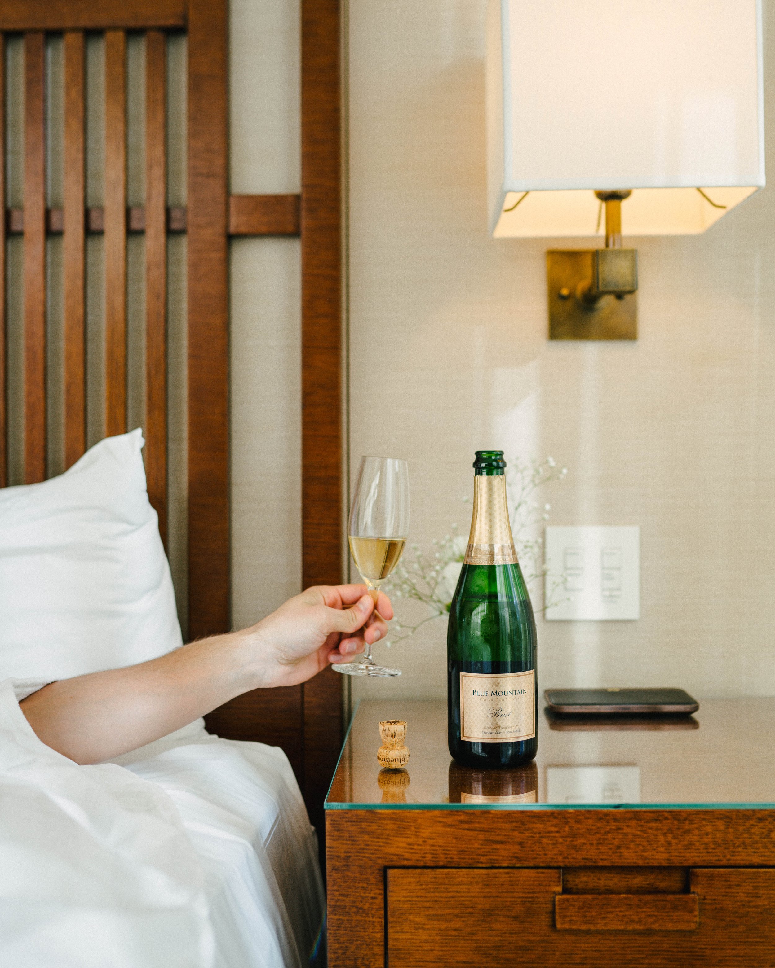 A person holding a glass of champagne next to a bottle of sparkling wine on a bedside table in a hotel room.