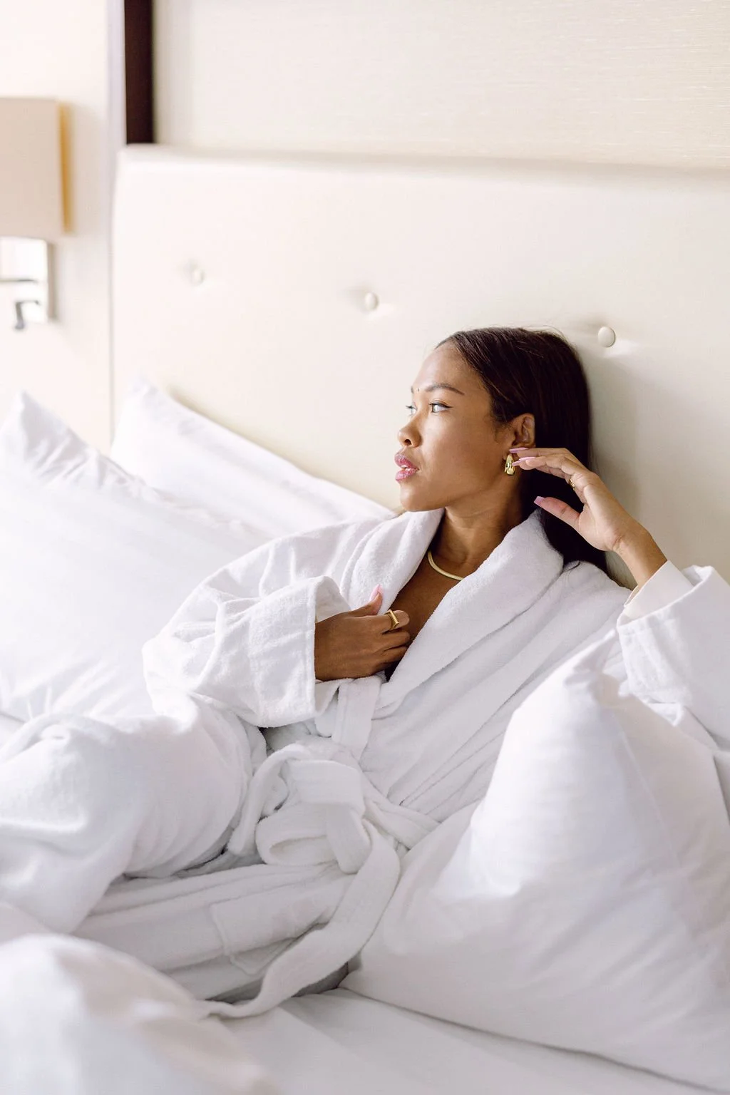 Woman in a white bathrobe sitting on a bed, touching her ear with one hand, looking to the side in a minimalist bedroom.