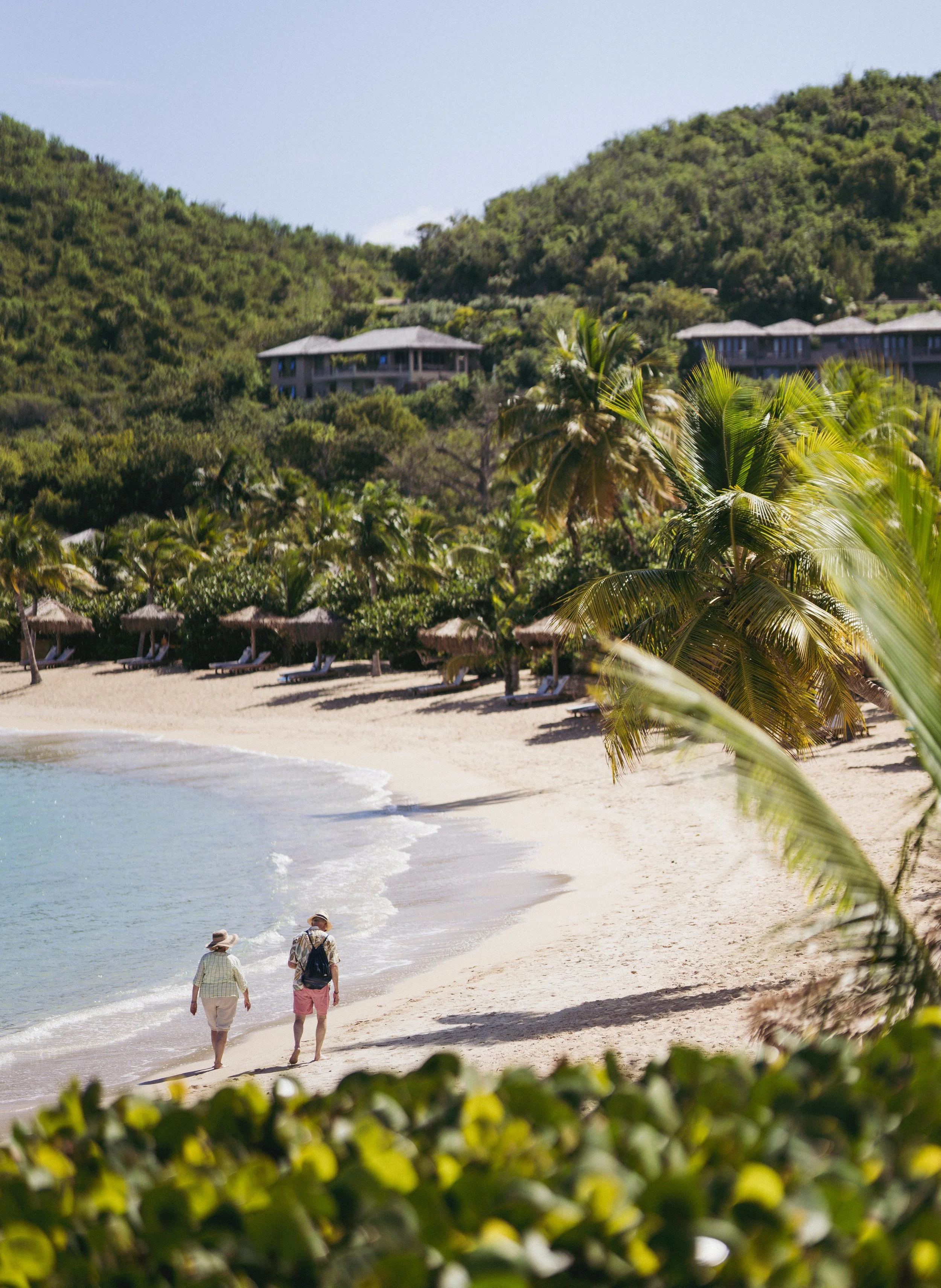 Two people walking along a sandy beach lined with palm trees and beach huts, with hills and houses in the background.