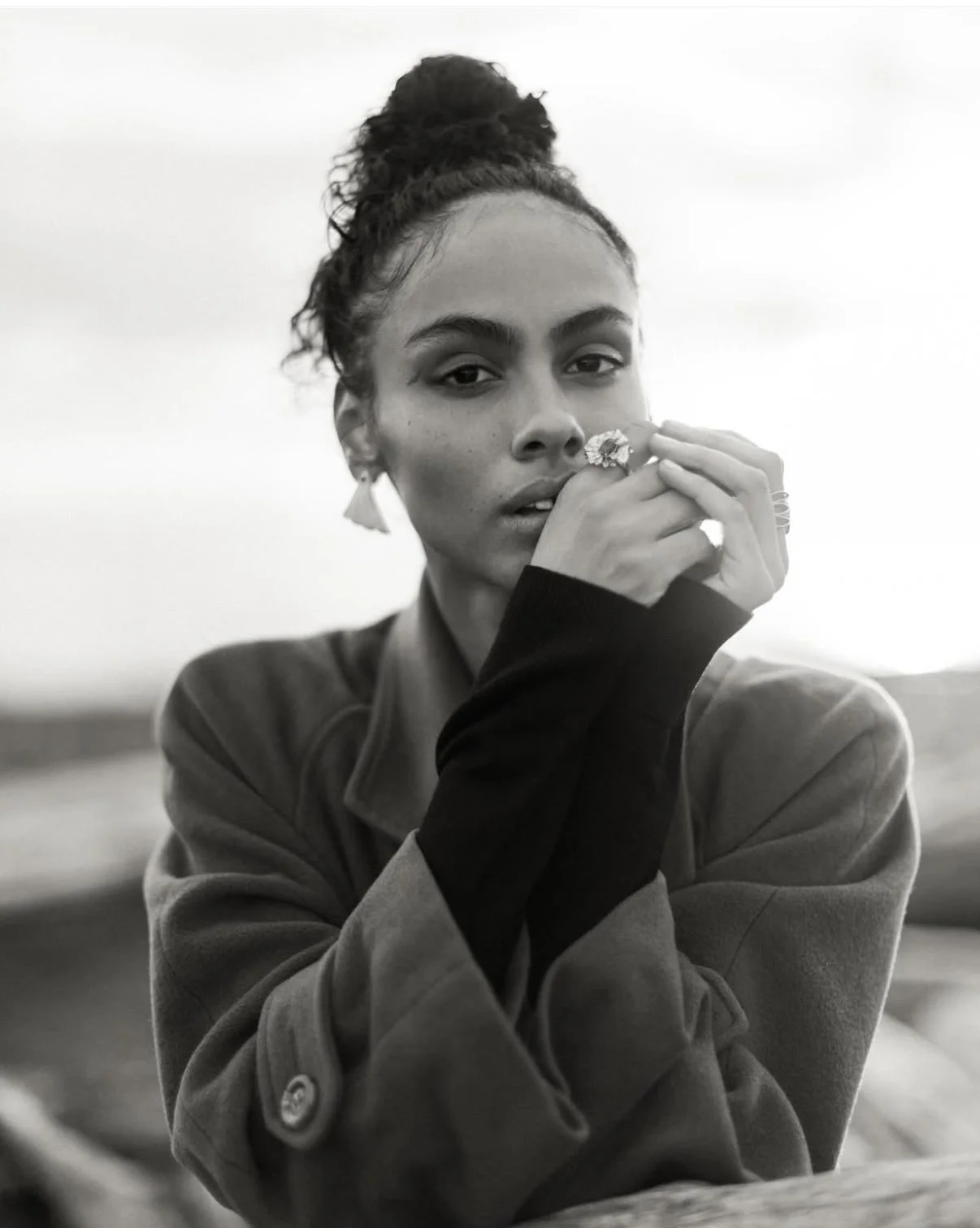 A young woman with dark hair in a bun, wearing a coat and rings, gazes at the camera in a black and white photo outdoors.
