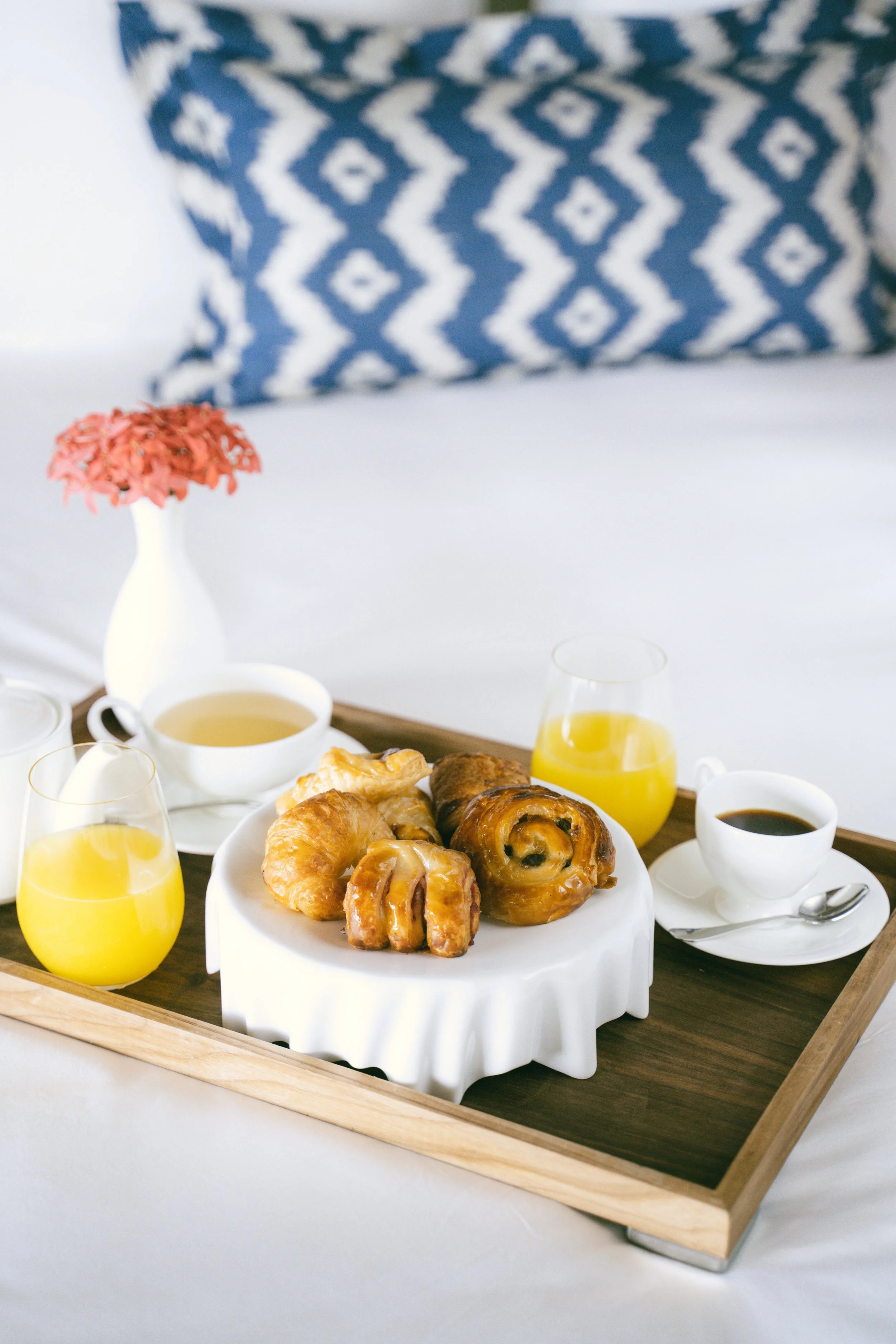 A breakfast tray with a white cake stand holding four croissants and cinnamon rolls, two glasses of orange juice, two cups of coffee, a cup of tea, a small vase with pink flowers, set on a bed with a blue and white patterned pillow in the background.