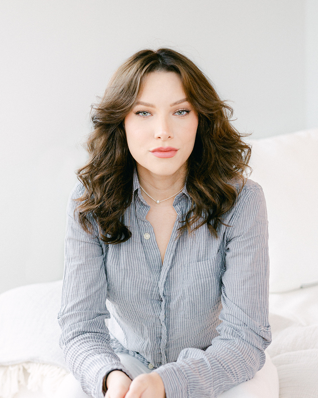 A woman with wavy brown hair and blue eyes wearing a striped button-up shirt and a necklace, sitting on a white couch in a bright room.