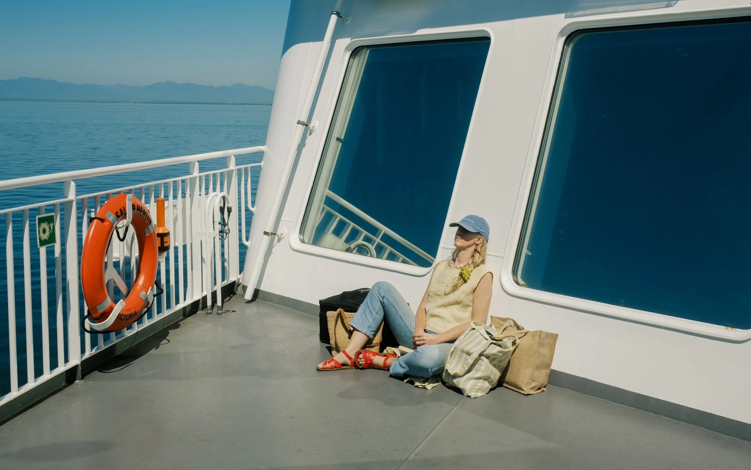 A woman relaxing on a boat deck, sitting against the white wall near large tinted windows, with water and mountains in the background, wearing a yellow top, blue jeans, red sandals, a blue cap, and sunglasses, with bags beside her and a life buoy hun